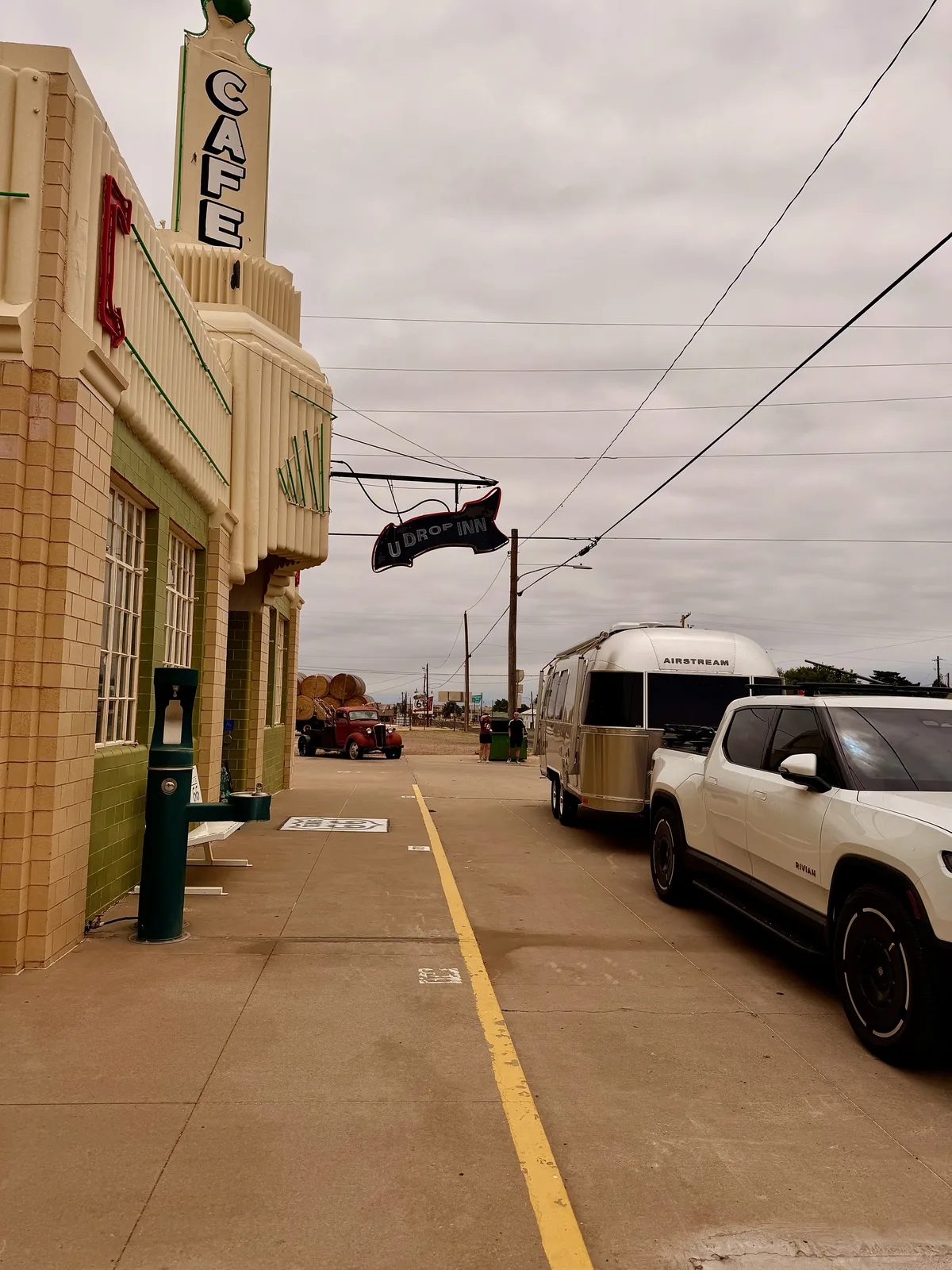 The U-Drop Inn from the street level, the cream Art Deco "CAFE" tower visible above and the Texas-shaped "U DROP INN" neon arrow sign hanging from a black bracket, the silver Airstream International + white Rivian R1T parked alongside, an antique red truck across the street for vintage Route 66 atmosphere