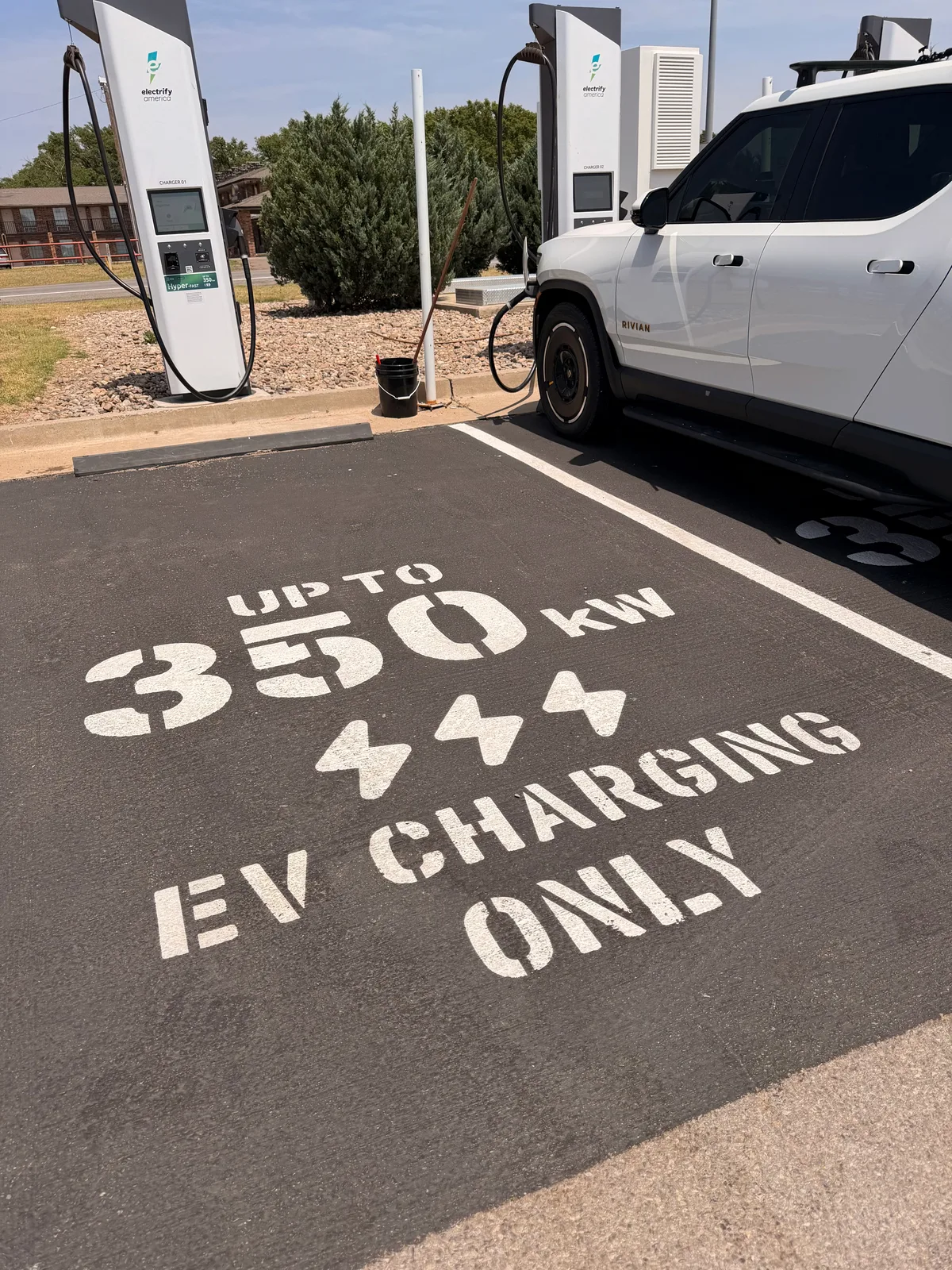Two Electrify America DC fast-charge pedestals at the Erick OK Love's Travel Stop with the white Rivian R1T plugged in, the asphalt in the foreground stenciled in white "UP TO 350 kW EV CHARGING ONLY", three lightning-bolt graphics between the digits and the text