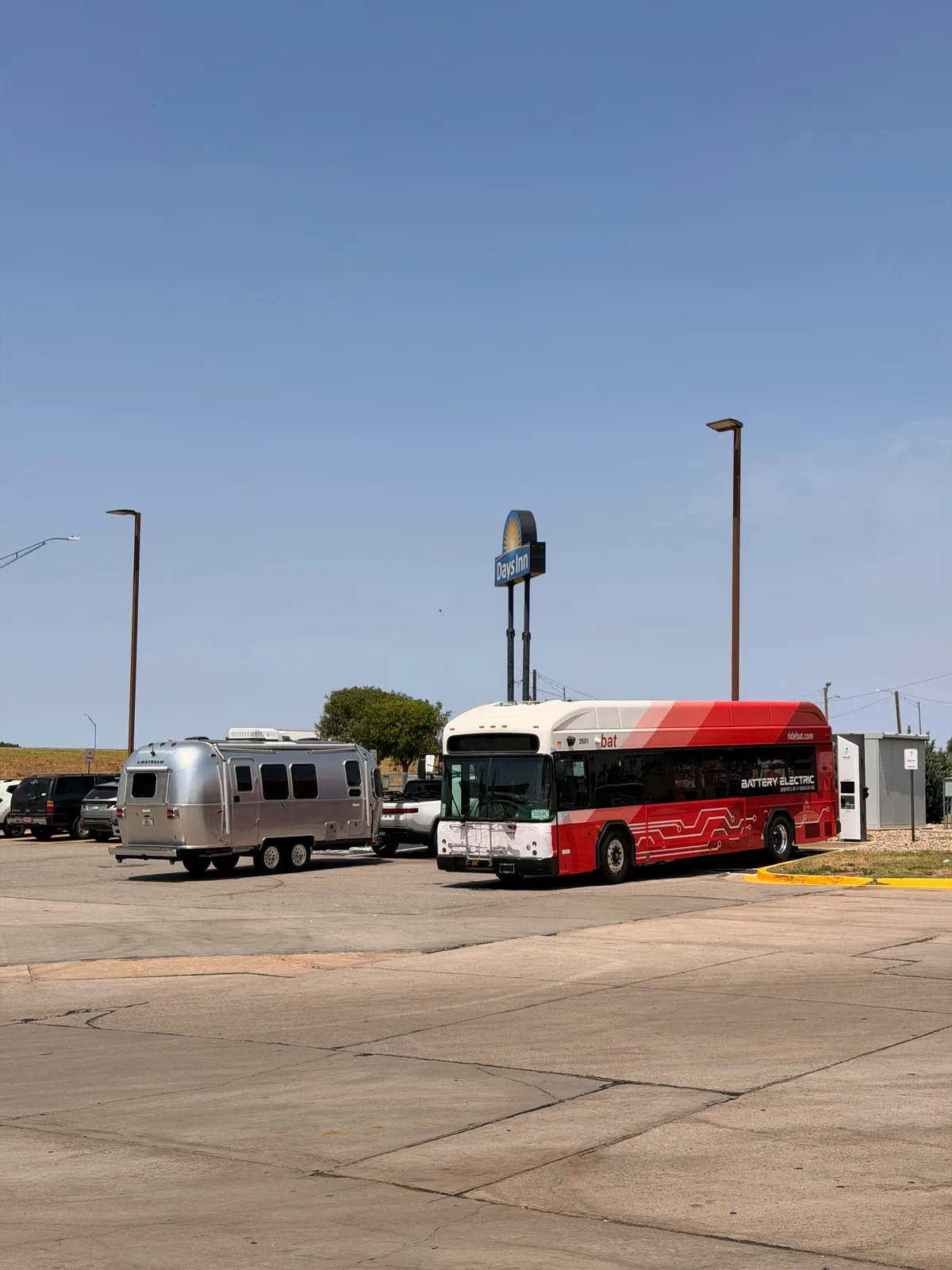The silver Airstream International parked next to a red-and-white battery-electric BAT regional transit bus at the Erick OK Love's Travel Stop, a Days Inn sign visible behind, two electric vehicles on the same lot