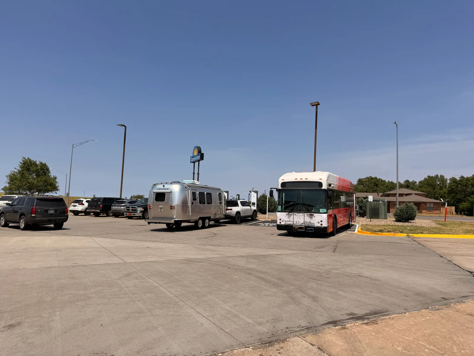 Wide-angle view of the Erick OK Electrify America charging lot, the silver Airstream and white Rivian R1T plugged in at left, a red battery-electric BAT bus to the right, a Days Inn motel sign rising behind under a clear blue Oklahoma sky