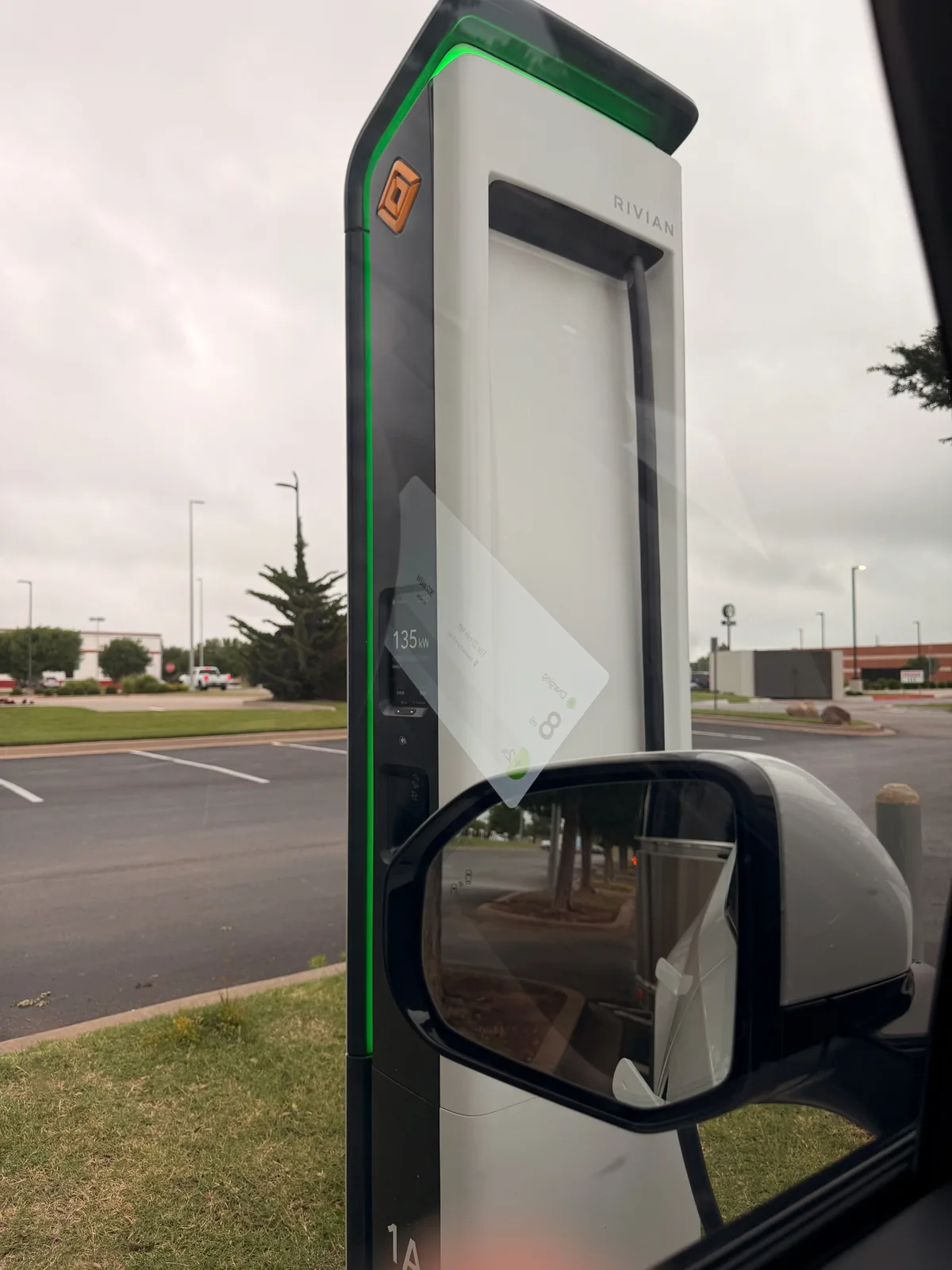 The tan-and-green Rivian Adventure Network pedestal 1A at the west-OKC RAN station, the Rivian wordmark and diamond R logo visible at the top, the pedestal display showing 135 kW current charge rate just after plug-in, the Rivian R1T side mirror in the foreground