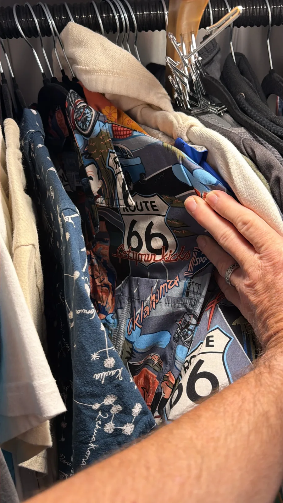 Inside the Airstream wardrobe at Rockwell RV Resort, a hand reaching for a hanging short-sleeve shirt with a Route 66 shield motif and "Oklahoma" lettering, surrounded by other Route 66 / Oklahoma themed shirts on hangers