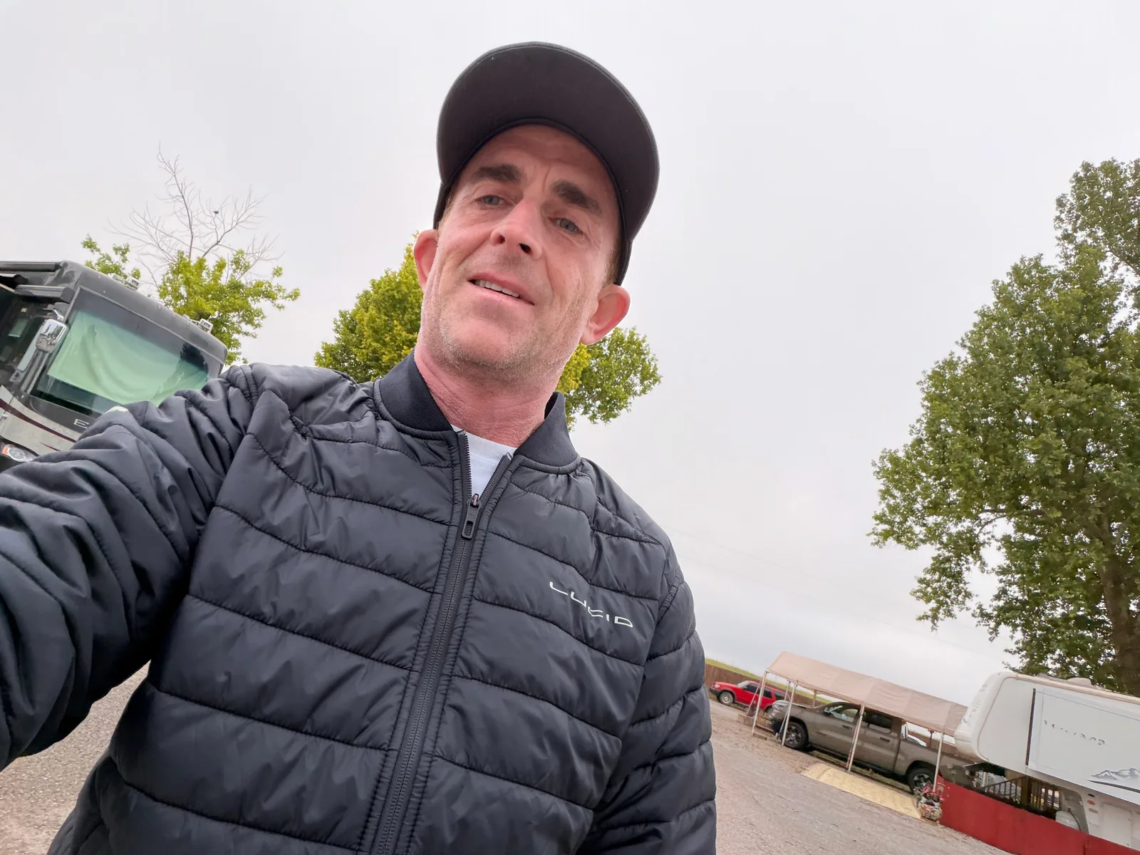 Eric in a black puffy LULU jacket and dark cap, selfie outside at Rockwell RV Resort with the white Rivian R1T side panel in the frame, a red pickup and white travel trailer across the lot, overcast Oklahoma sky