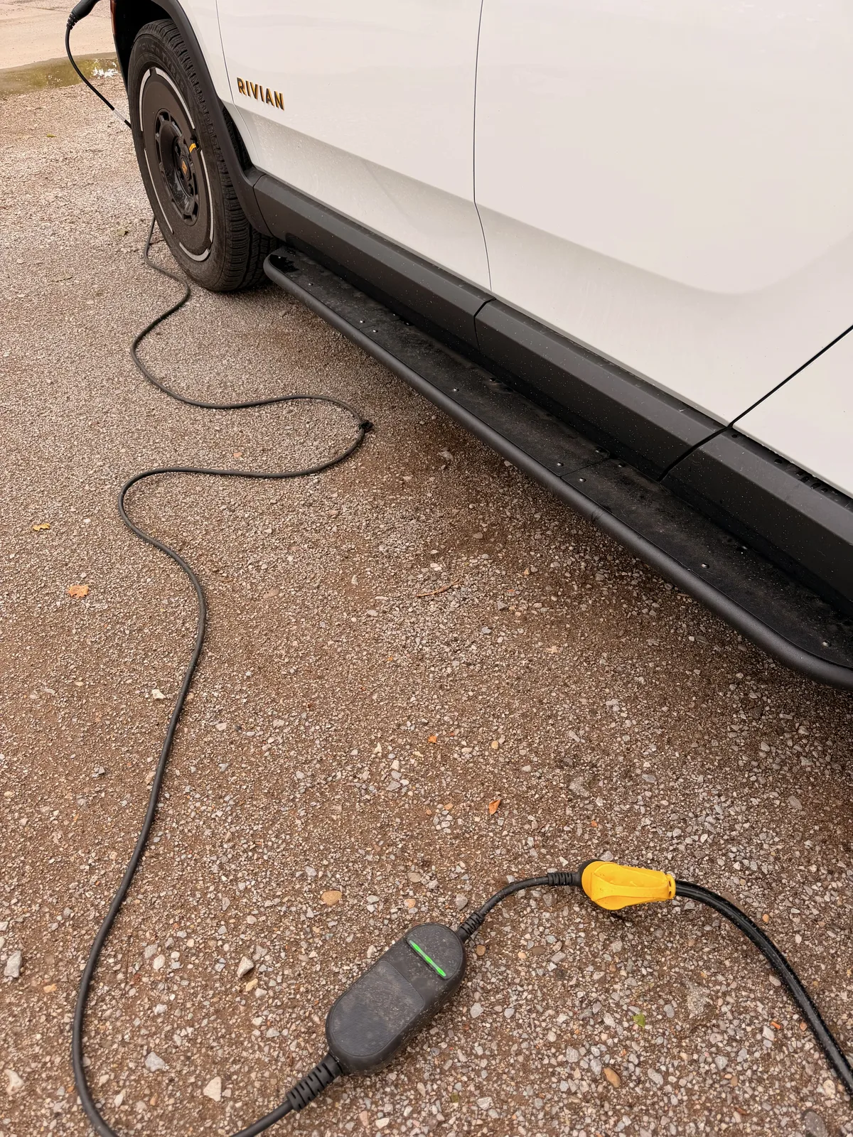 The white Rivian R1T rear quarter with "RIVIAN" wordmark on the side panel, the included Rivian portable charge cable snaking from the truck's NACS port across the gravel pad to the yellow NEMA 14-50 head plugged into the 50A pedestal at Rockwell RV Resort