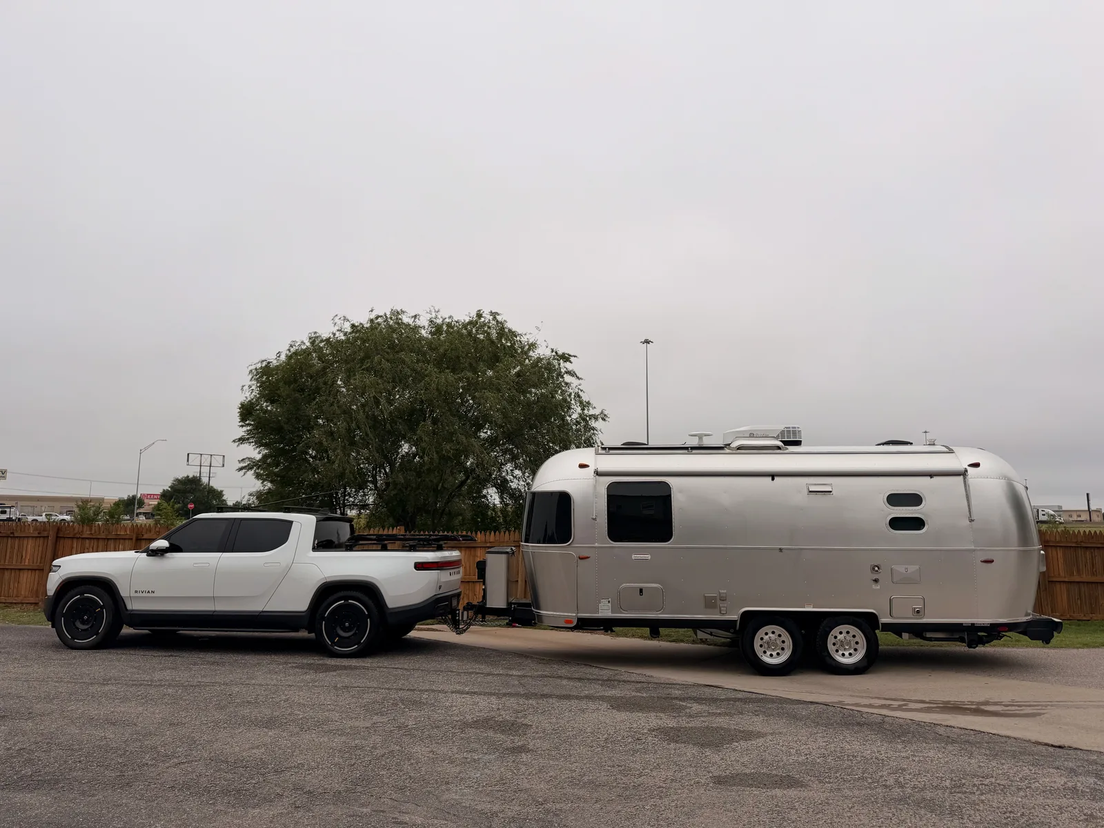 The white Rivian R1T hitched to the silver Airstream International parked broadside in the Rockwell RV Resort lot, a tree at left center, an overcast Oklahoma plains sky, ready to roll