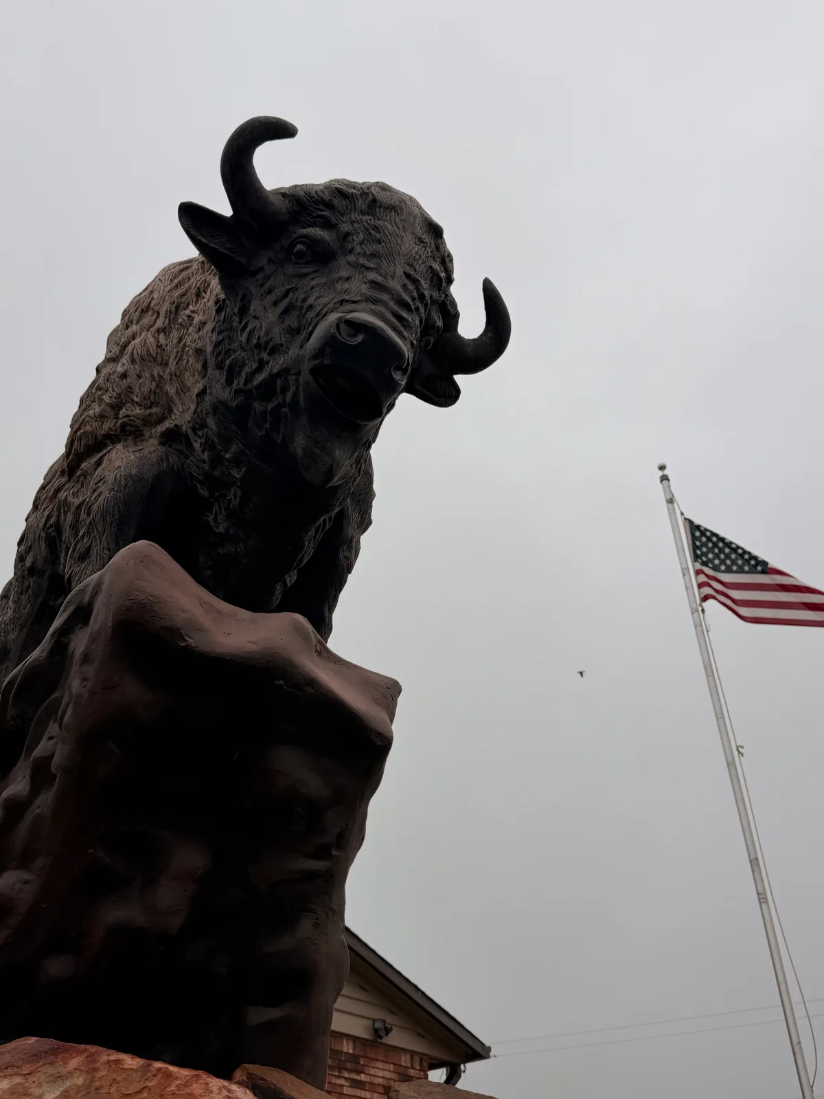 A dark-bronze bison sculpture mounted on a rust-red rock base near the Rockwell RV Resort entrance with the head tilted up to the left, a tall American flag on a white pole at right, an overcast Oklahoma plains sky