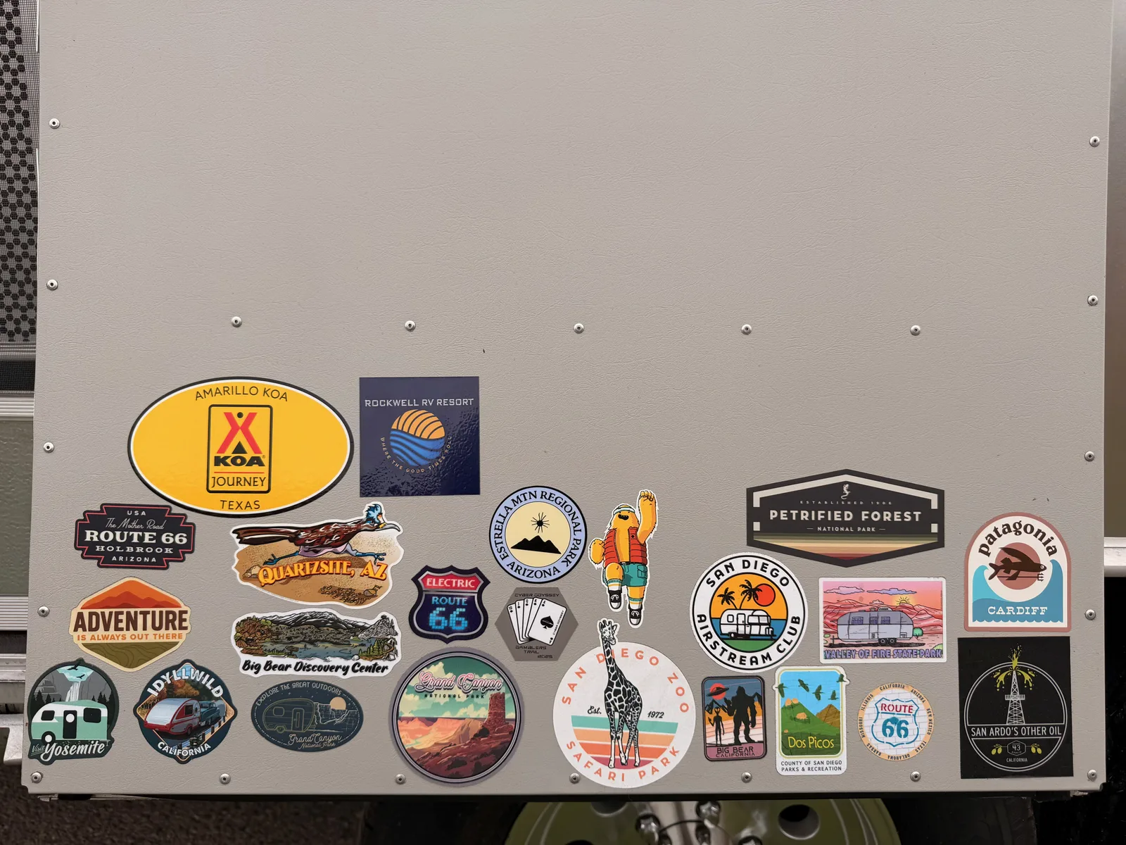 The aluminum-finished sticker panel mounted on the side of the Airstream International, covered in collected stickers from previous trip stops including Amarillo KOA Texas, Route 66 Holbrook Arizona, Petrified Forest National Park, Big Bear Discovery Center, San Diego Airstream Club, Patagonia Cardiff, Yosemite, San Diego Zoo Safari Park, the Electric Route 66 logo, plus a fresh round blue Rockwell RV Resort badge just added