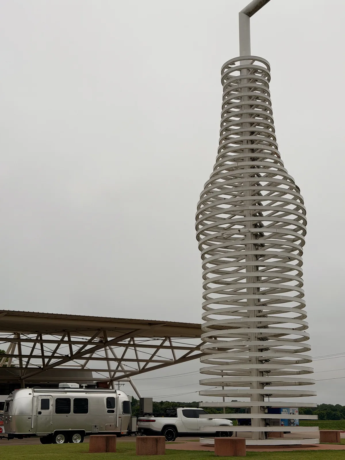 Vertical view of the 66-foot Pops 66 stainless steel bottle sculpture rising into the overcast sky with the architectural canopy and the silver Airstream visible at the base, a slight angle variant from the primary hero shot