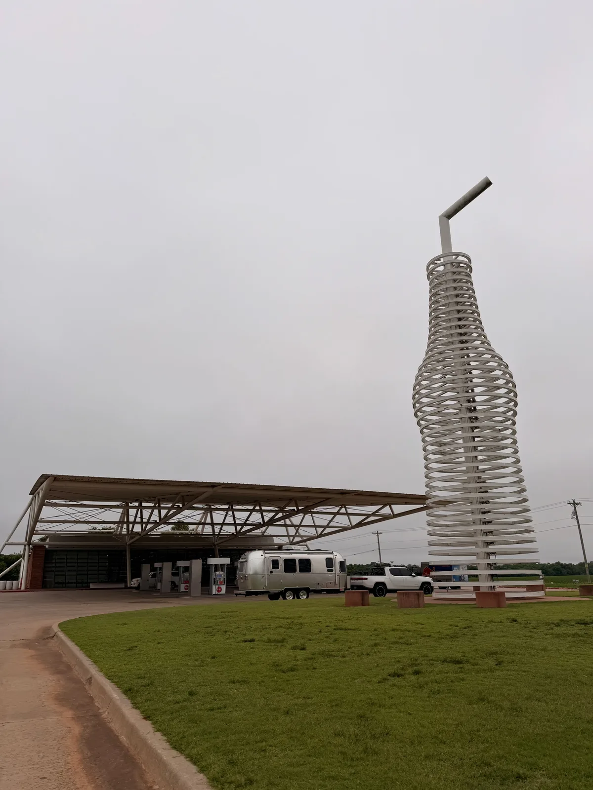 Wide-angle view of Pops 66 Soda Ranch in Arcadia OK with the full 66-foot pop bottle sculpture at right, the sweeping architectural canopy over the gas and charging area at left, the silver Airstream International and white Rivian R1T parked under the canopy mid-frame, overcast Oklahoma plains sky