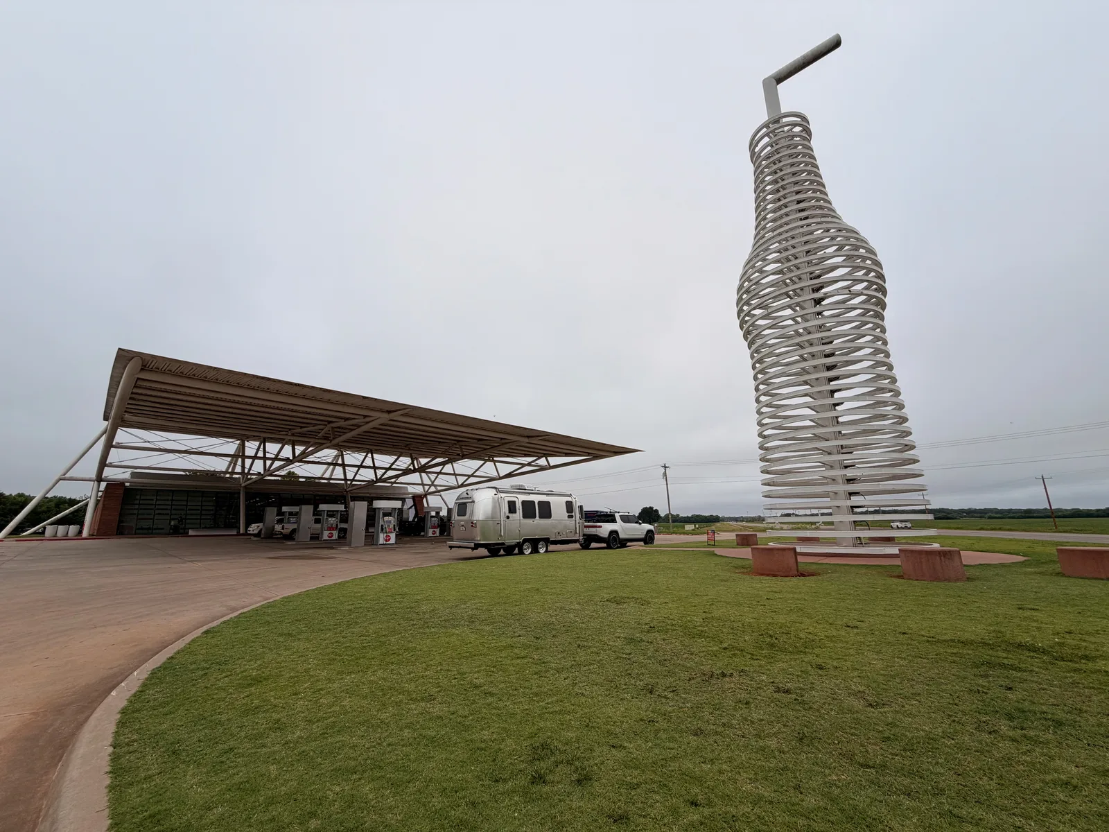 Wide ground-level view of Pops 66 Soda Ranch from the front lawn corner, the swept architectural canopy at left running across the gas + charging bays, the 66-foot stainless steel bottle sculpture at right rising into the overcast sky, the silver Airstream and white Rivian R1T parked between, grass apron and bench seating in the foreground