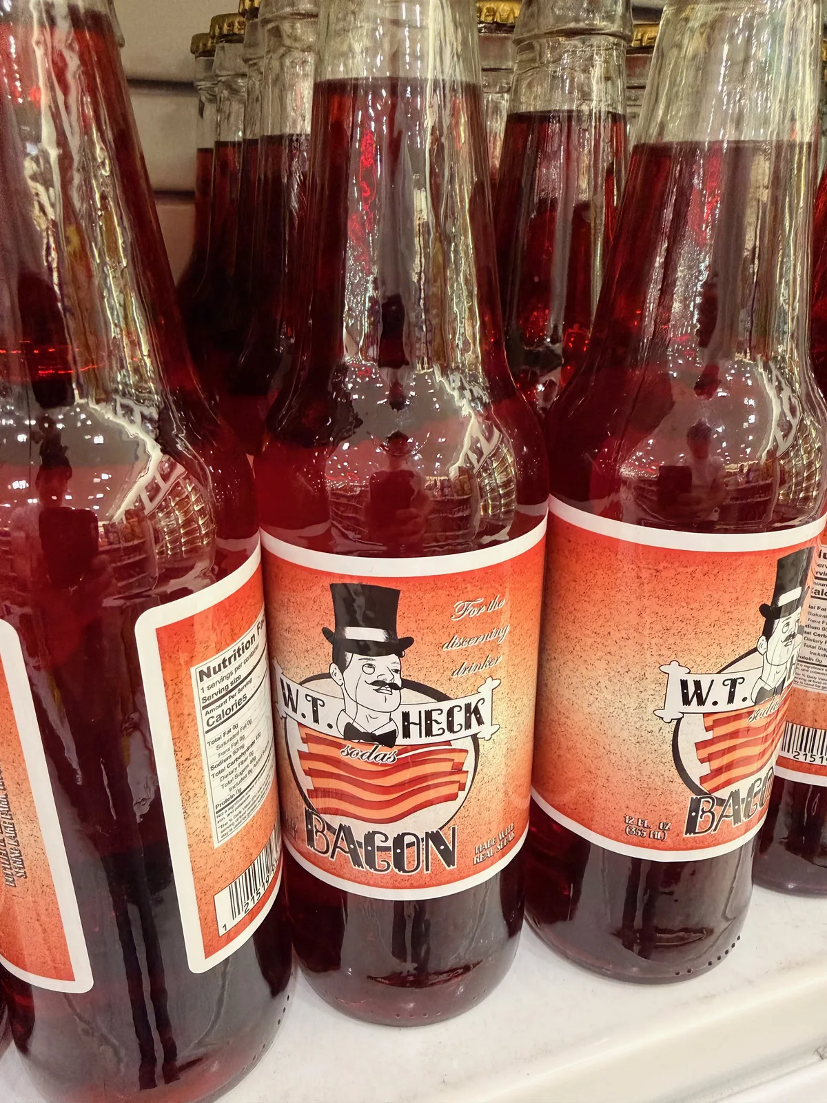Close-up of a W.T. Heck Bacon Soda bottle on the Pops 66 shelf with a top-hat-and-mustache mascot illustration on a label of bacon stripes, ruby-red liquid visible through the clear glass, identical bottles flanking it