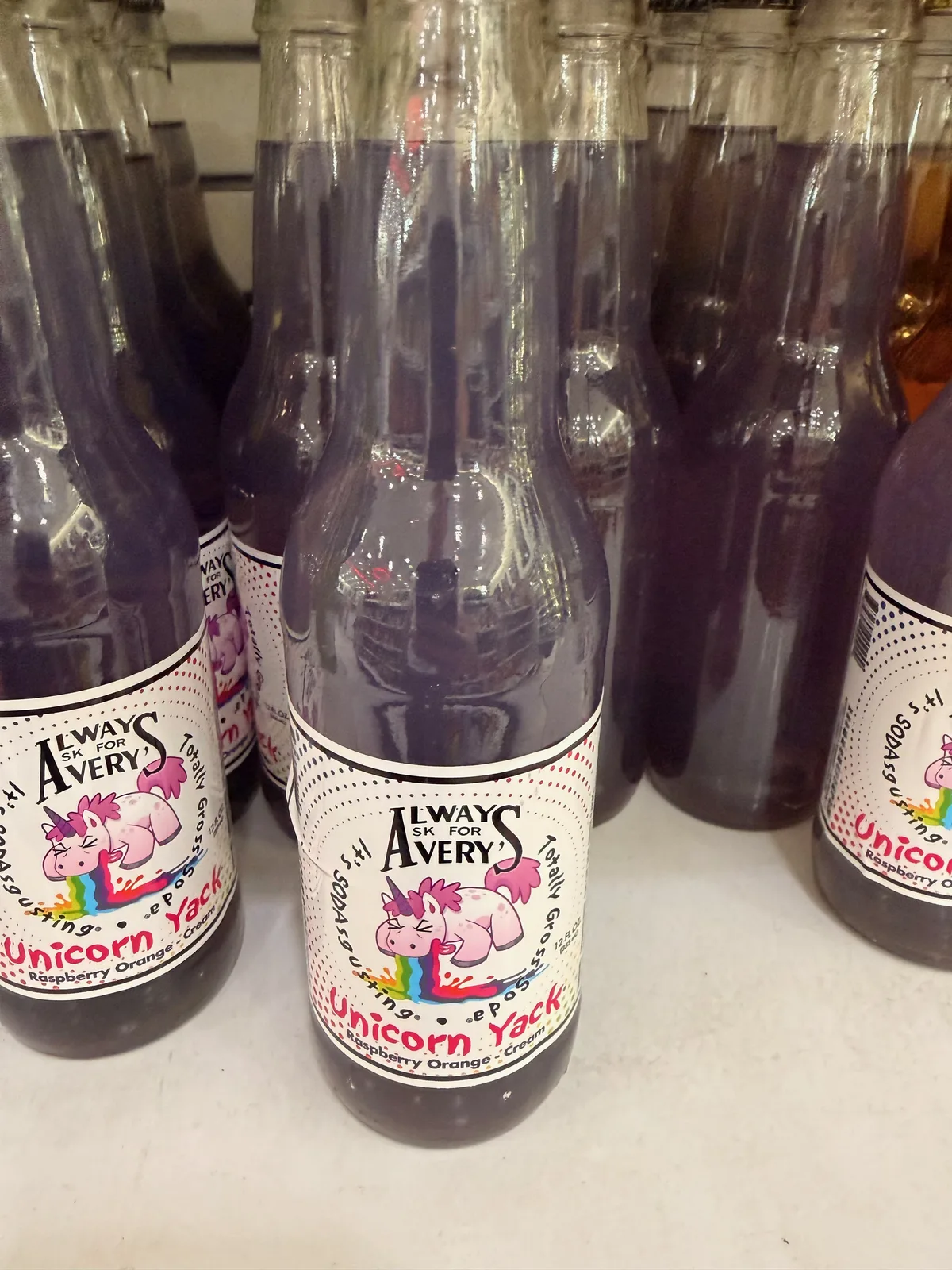 Close-up of an Always Ask for Avery's Unicorn Yack soda bottle at Pops 66 with a cartoon pink unicorn on the label, "raspberry orange cane sugar" caption, purple-tinted liquid in clear glass, more identical bottles behind