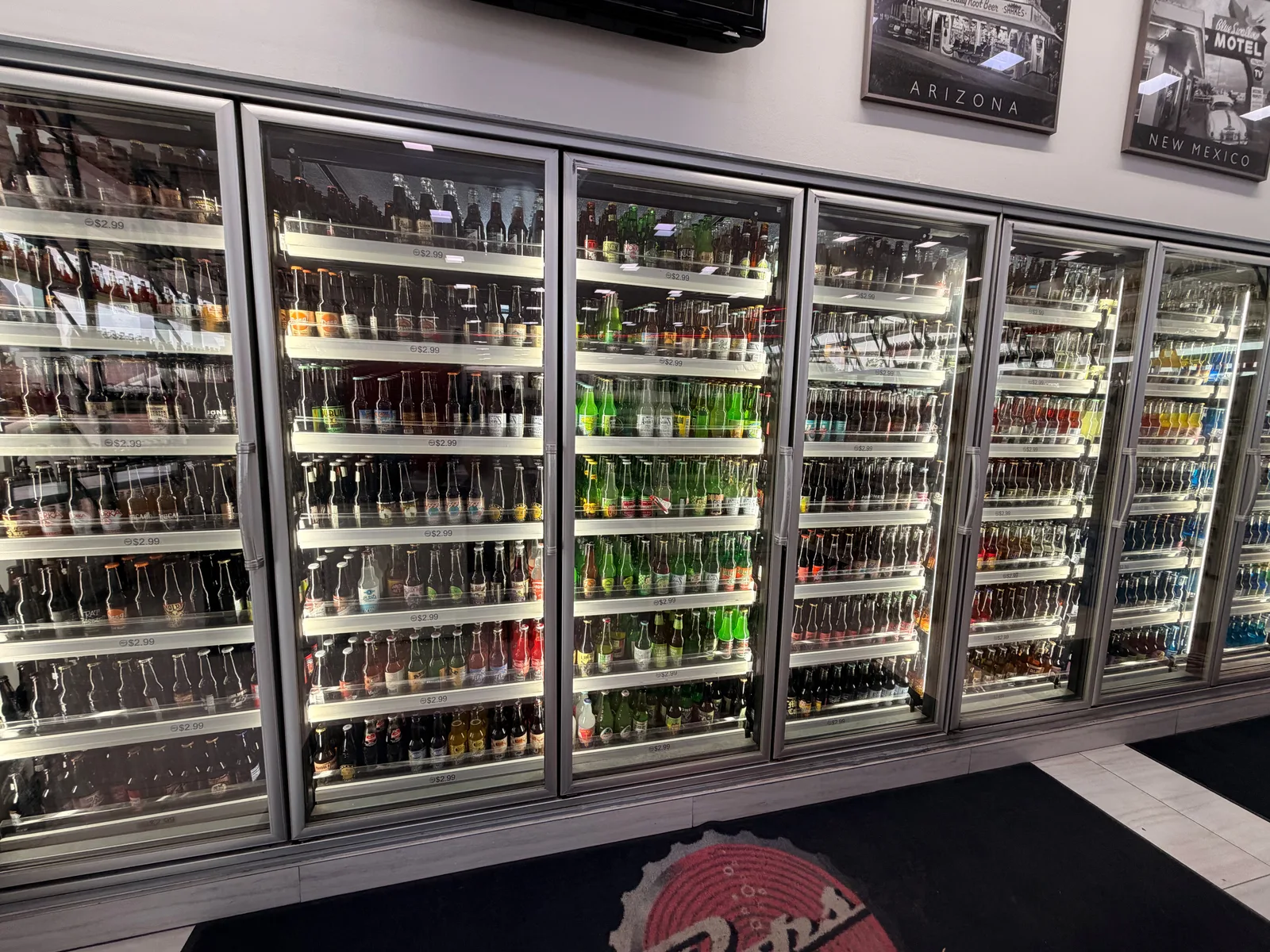 The Pops 66 wall of glass-front soda coolers running floor-to-ceiling with hundreds of bottles arranged by color and label, ARIZONA and NEW MEXICO Route 66 photo panels mounted above the cases, the Pops 66 floor logo visible at bottom right