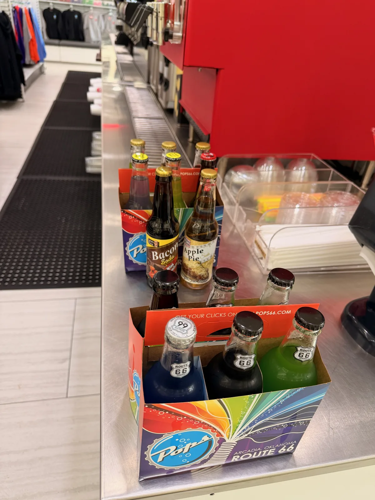 Inside the Pops 66 gift-shop at the checkout counter, a Pops 66 four-pack box with blue, black, and green Route-66-branded soda bottles in front, single bottles of Bacon Soda and Apple Pie soda on the counter behind, retail apparel racks visible in the background