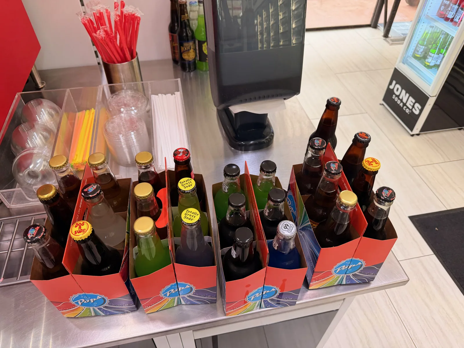 Five Pops 66 four-pack boxes lined up on the checkout counter, each with rainbow Route 66 themed graphics, filled with assorted glass soda bottles in red, green, blue, and brown colors with bottle caps still on, a Jones Soda glass-front cooler in the background