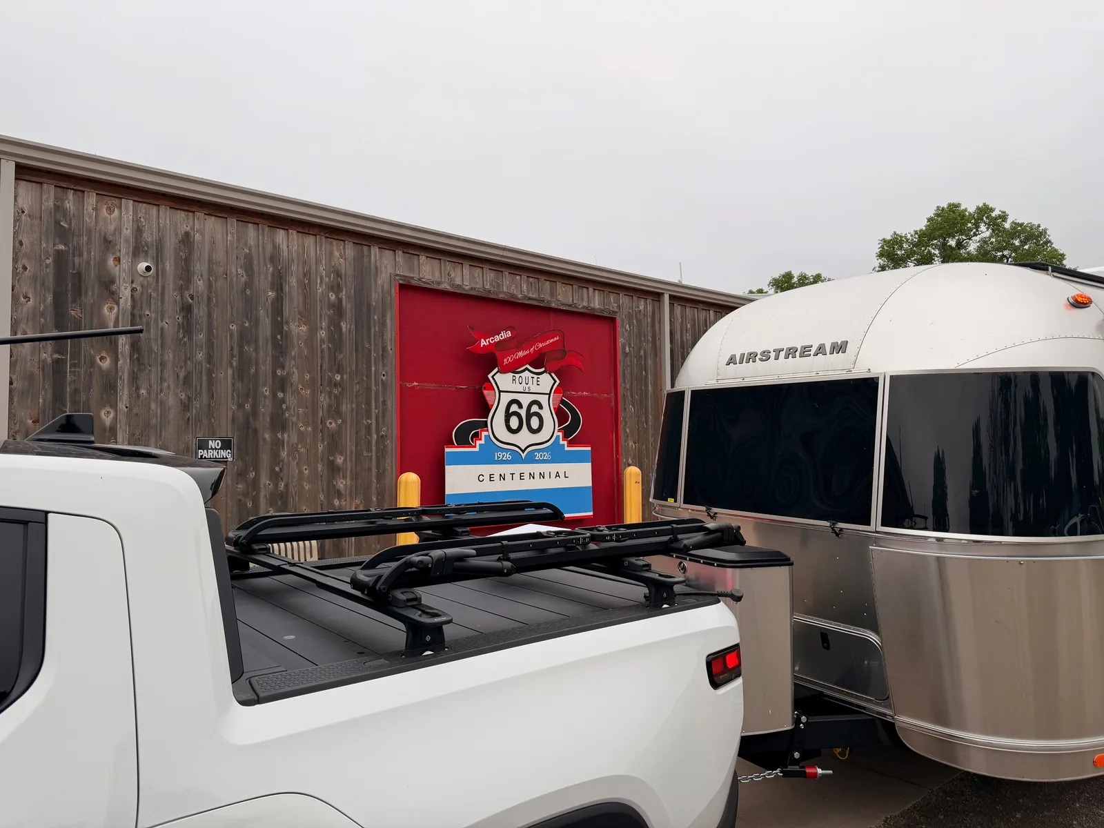 The white Rivian R1T tailgate with its black cross-rack at left foreground, the silver Airstream International rear at right, framing a bright red roll-up door on a wood-sided Arcadia building that holds a large black and white Route 66 shield panel with "1926 - 2026" centennial dates and CENTENNIAL on a blue-and-white banner below, an overcast Oklahoma sky above