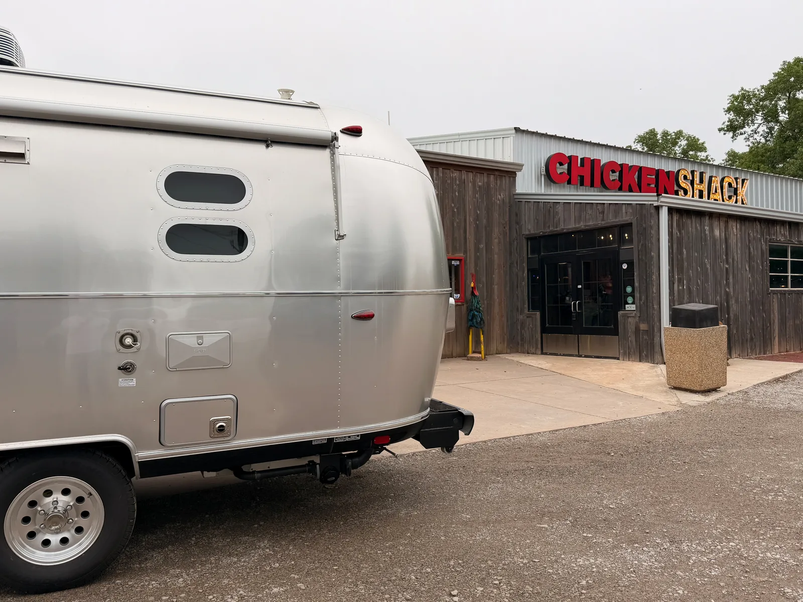 The silver Airstream International side panel at left foreground, the wood-sided Chicken Shack restaurant entrance at right with bold red-and-yellow "CHICKEN SHACK" block-letter signage above the doorway, a parking lot bollard and a trash receptacle by the entrance, overcast plains sky