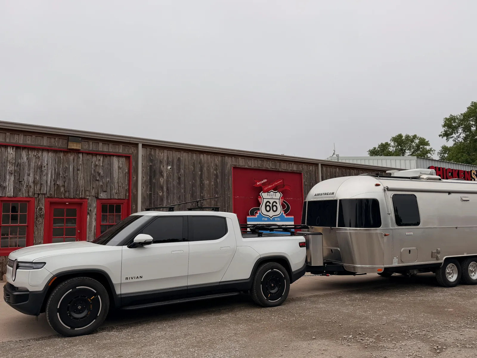 Wide view of the Arcadia Route 66 plaza, the white Rivian R1T and silver Airstream parked along the gravel lot, the wood-sided Chicken Shack / Centennial sign building running across the back, red roll-up doors and the centennial panel mid-frame, the partial "HILL'S" signage visible at right
