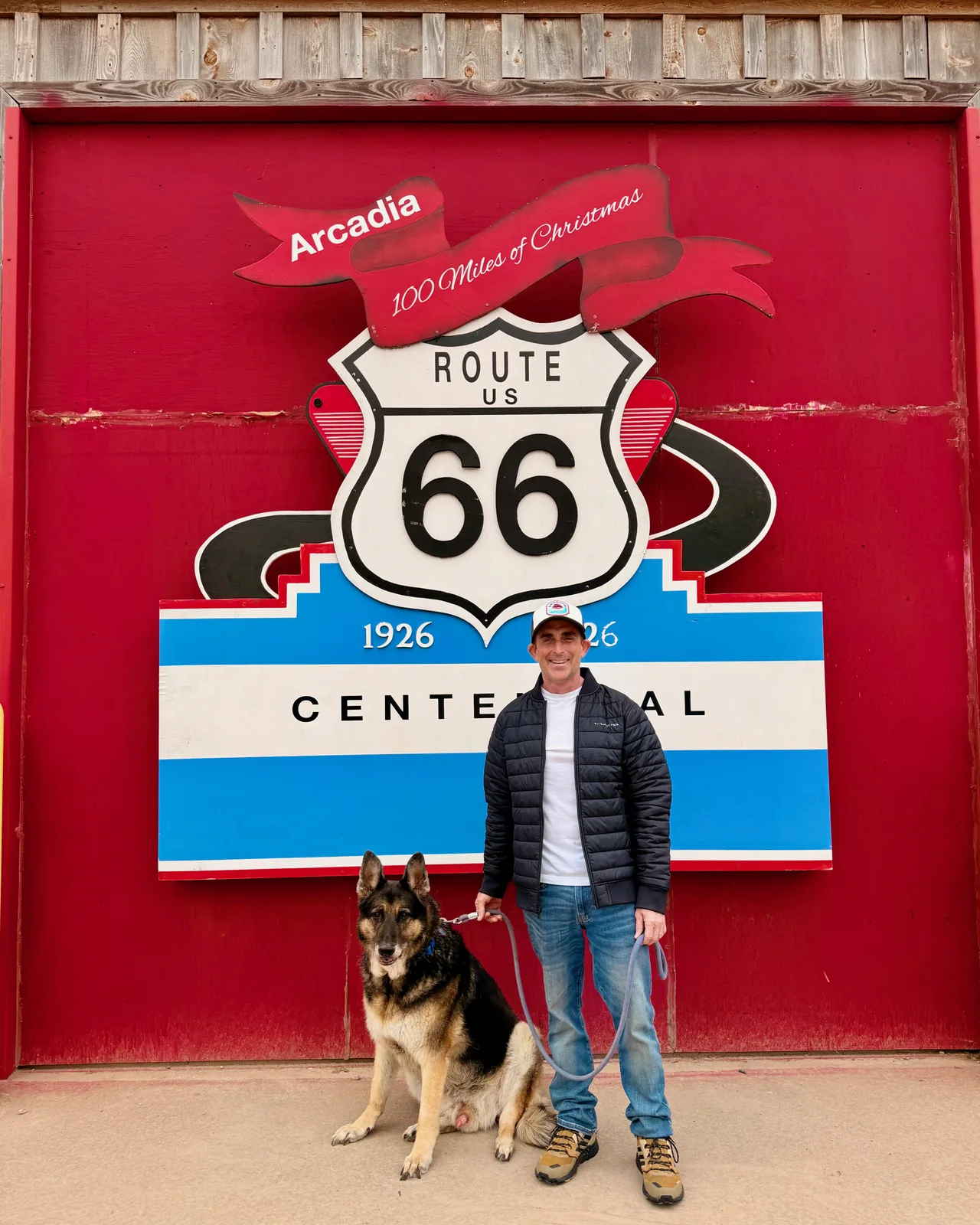 Eric in a black puffy LULU jacket and white cap standing in front of the Arcadia Route 66 Centennial sign, the family German Shepherd Mission sitting at his feet to the left looking right with leash in hand, the banner above reading "Arcadia" and "100 Miles of Christmas", the Route 66 1926 to 2026 Centennial panel filling the red roll-up door behind