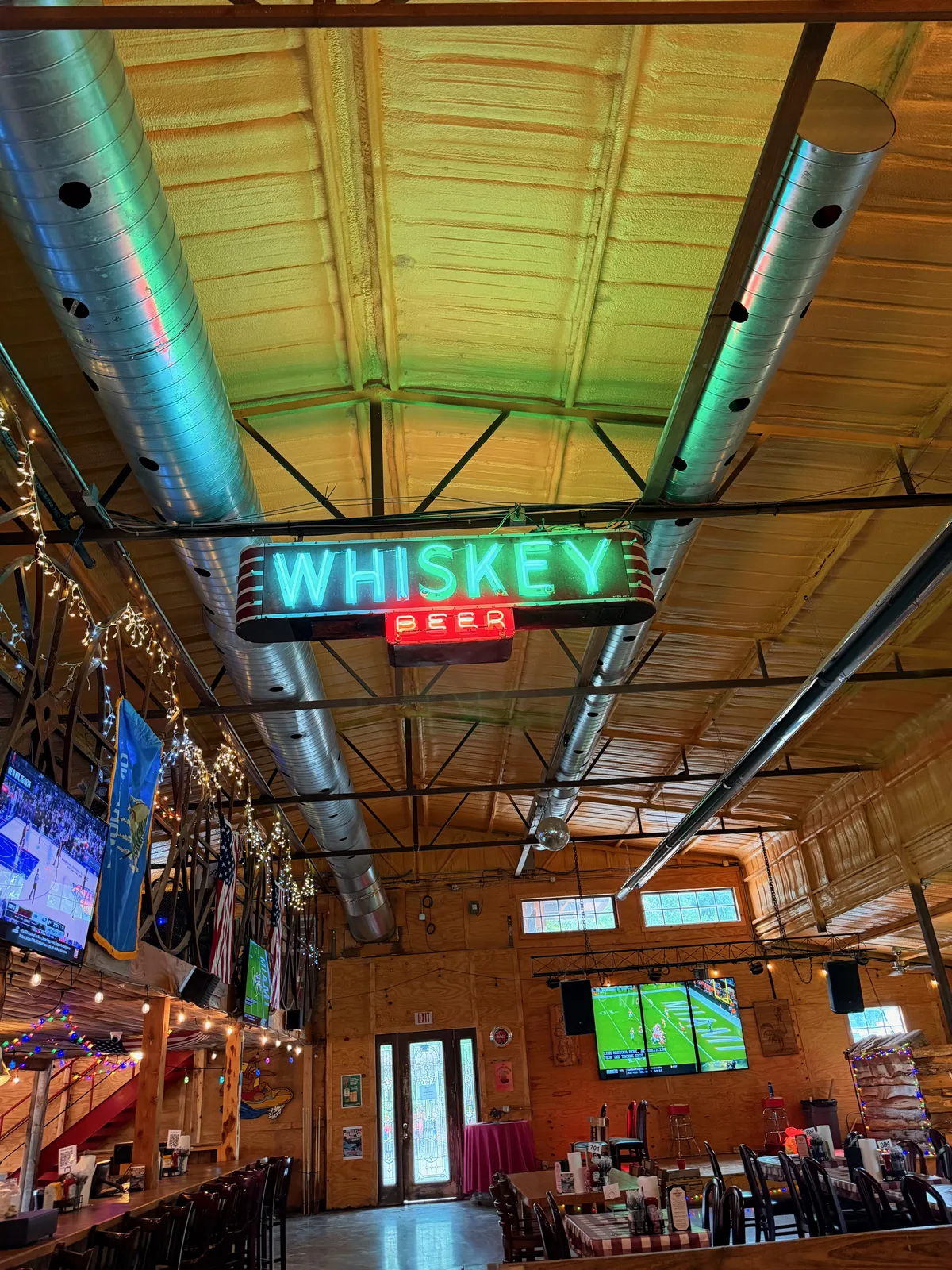 Inside the Chicken Shack restaurant looking up at the rafters, a "WHISKEY / BEER" neon sign hanging from a corrugated metal ceiling with industrial steel duct work, exposed brick walls below with multiple flat-panel TVs showing sports, string lights and Route 66 themed decor, dining tables visible at right