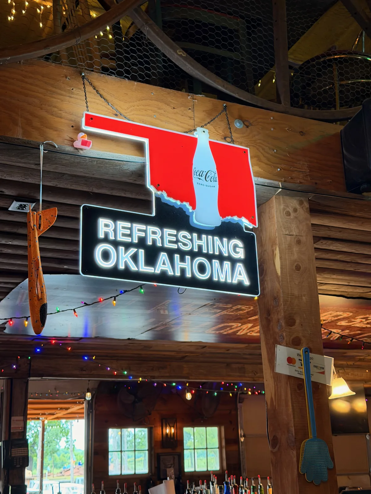 An Oklahoma-shaped Coca-Cola neon sign hanging from the wooden rafters of the Chicken Shack interior, a stylized white Coca-Cola contour bottle on a red Oklahoma silhouette with neon "REFRESHING OKLAHOMA" lettering beneath, multicolored string lights below, wooden support beams flanking