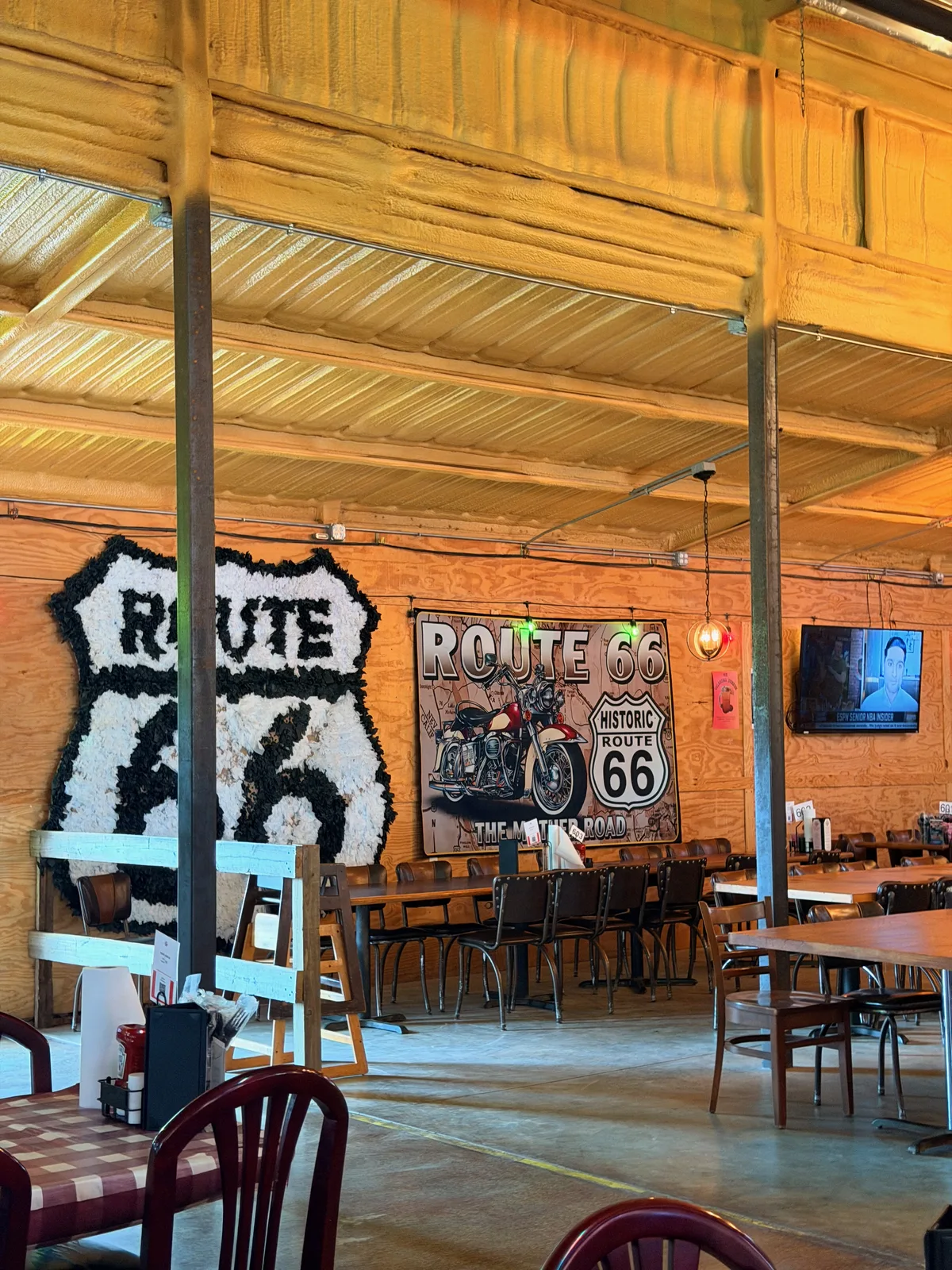 Interior dining area of the Chicken Shack with a large black and white floral Route 66 shield wreath on a wood-panel wall at left, a metal "Route 66 / Historic Route 66 / The Mother Road" tin sign with a motorcycle illustration at center, wood booth seating and red-checkered tablecloths in the foreground, corrugated metal ceiling above
