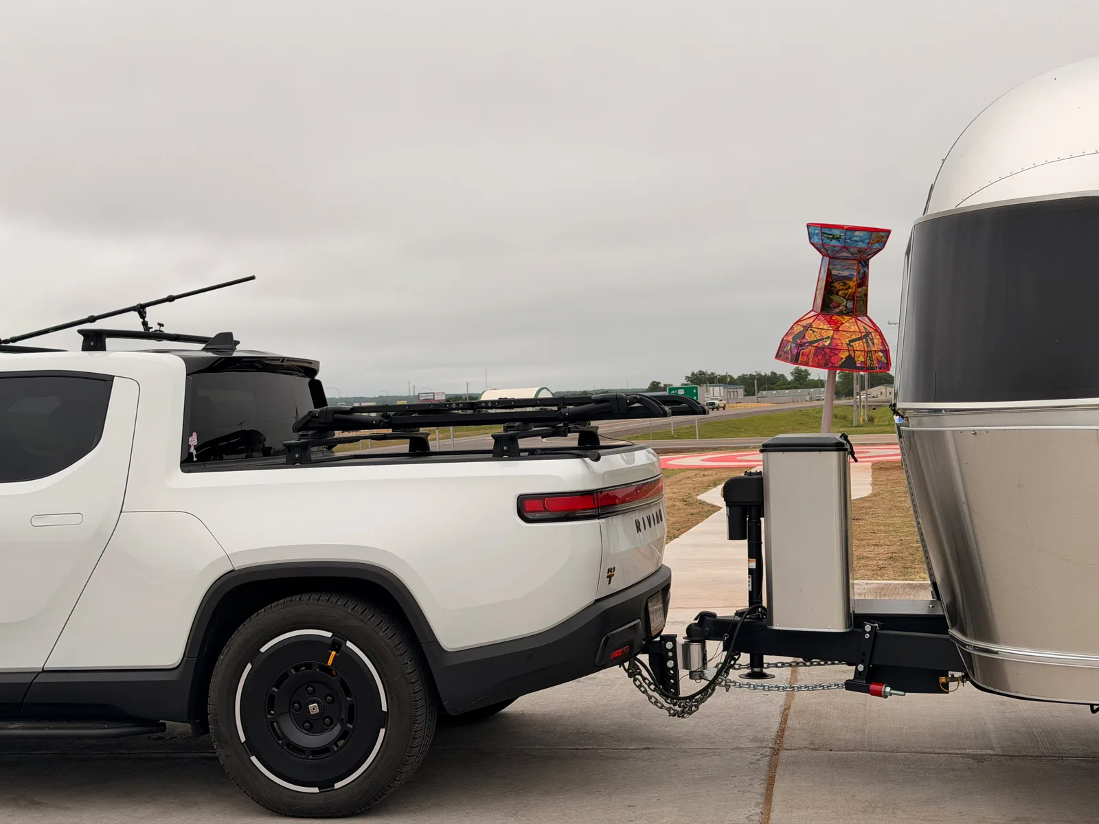 The white Rivian R1T hitched to the silver Airstream International at a roadside pull-off on old Route 66 east of Arcadia OK, with a tall mosaic stained-glass art totem in vivid patchwork color standing beside the trailer, overcast plains sky and a red-curbed turnout