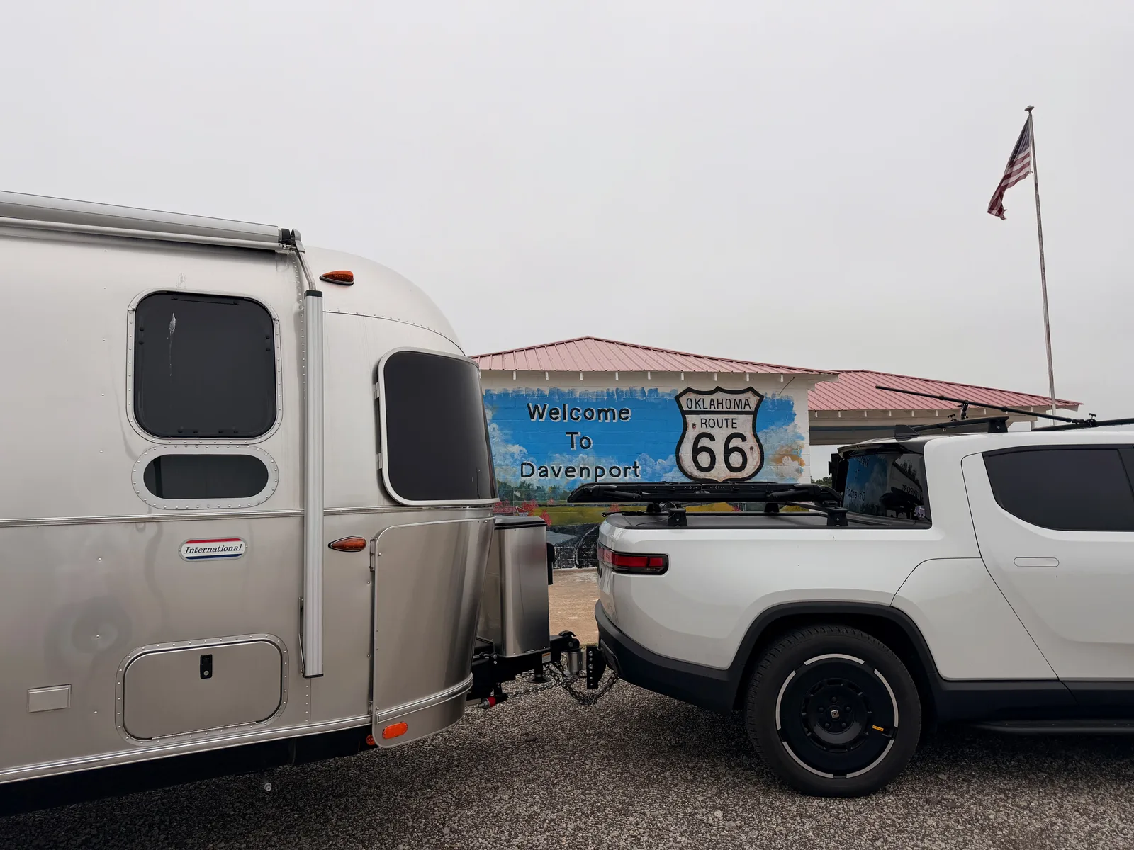The silver Airstream and white Rivian R1T parked in front of a "Welcome To Davenport" Route 66 mural building in Davenport OK, the Oklahoma Route 66 shield painted at right, an American flag flying above the metal-roofed structure