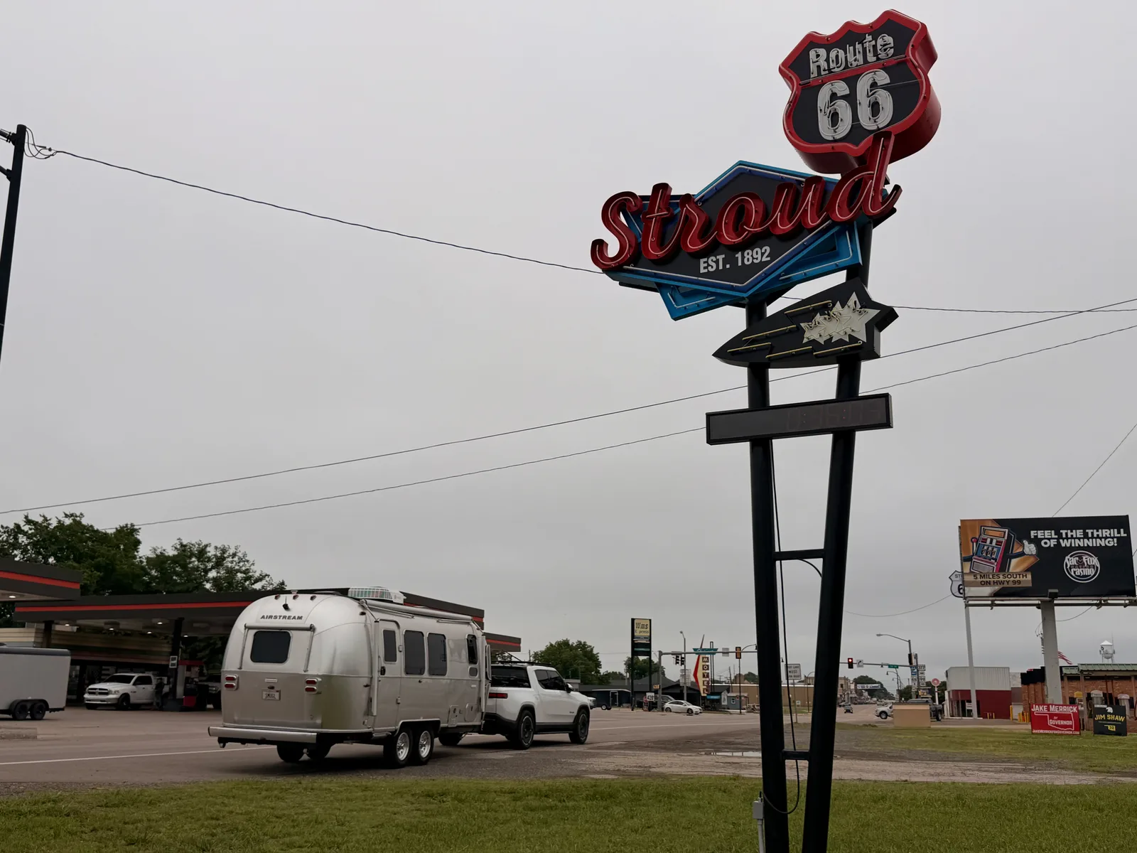 The red-and-blue "Route 66 Stroud EST. 1892" neon sign on a tall pole over the main drag in Stroud OK, the Route 66 shield at top and Stroud in script below, the silver Airstream and white Rivian R1T passing on the road behind