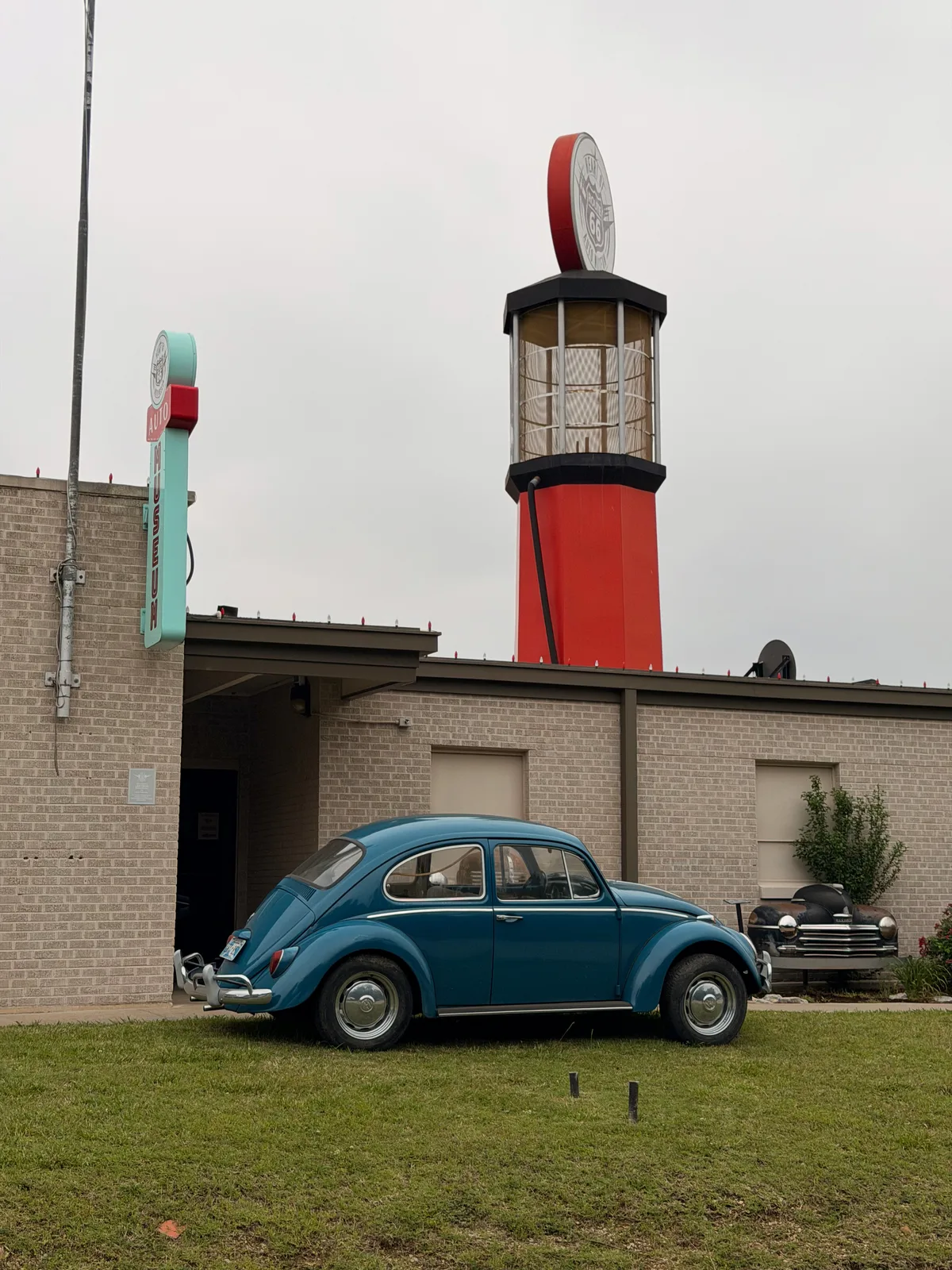 A vintage blue Volkswagen Beetle parked in front of the Heart of Route 66 Auto Museum in Sapulpa OK, with the museum's 66-foot-tall stylized red gas-pump tower rising behind topped by a clock-faced globe, a 1940s sedan parked to the right