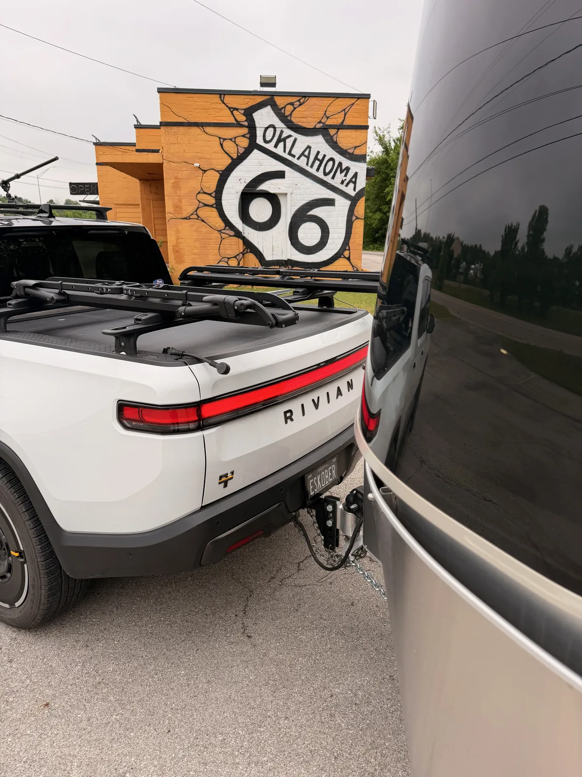 A large OKLAHOMA Route 66 shield mural painted on a cracked-plaster brick wall at the Heart of Route 66 Auto Museum in Sapulpa, framed by the white Rivian R1T rear with the SMUGL plate at left and the curve of the silver Airstream at right