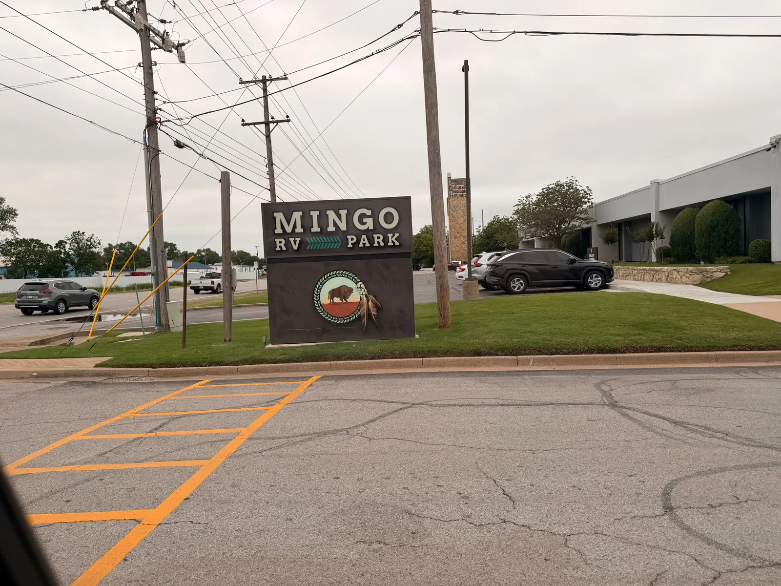The Mingo RV Park entrance monument sign in Tulsa, dark stone with "MINGO RV PARK" in white letters above a round tribal-medallion logo, set in a grass median with power lines and a low office building behind under an overcast sky