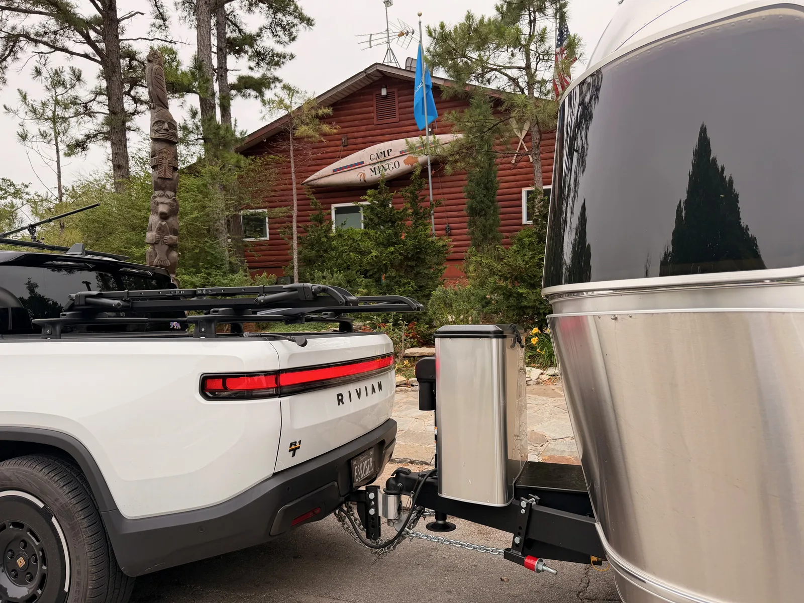The white Rivian R1T and silver Airstream parked at a Mingo RV Park site in Tulsa, with the red log-cabin Camp Mingo office building behind among pine trees, the Oklahoma state flag flying and a tall carved totem pole at left