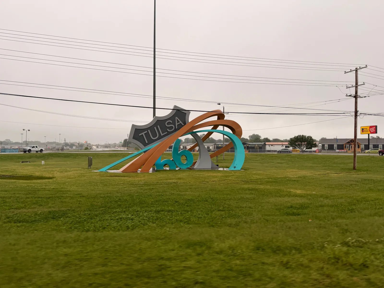 The Route 66 Rising sculpture at the Avery Traffic Circle in Tulsa, a large steel Route 66 shield reading "TULSA" with an oversized "66" half-submerged in a grassy island and ringed in swirls of tan, orange, and teal, under an overcast sky