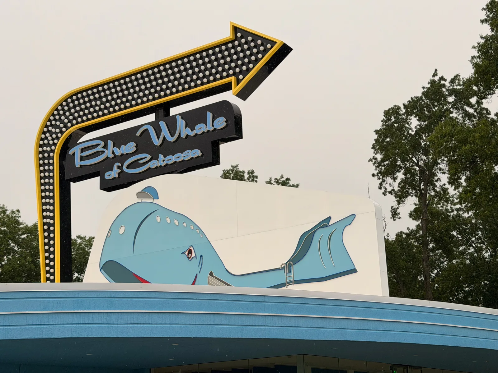 The "Blue Whale of Catoosa" sign at the attraction entrance, a black starburst-bordered panel with a painted leaping light-blue whale and yellow arrow, mounted above the pond's edge with green trees behind under a grey sky