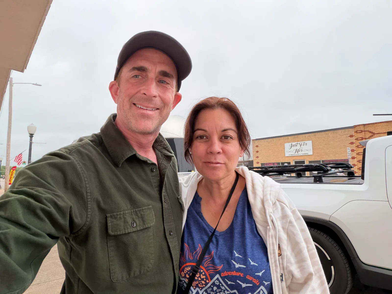 Eric in an olive shirt and dark cap and Diana in a blue tee and cream cardigan, selfie outside Hometown Coffee on the main street of Locust Grove OK, the white Rivian R1T and a brick storefront behind them, overcast morning