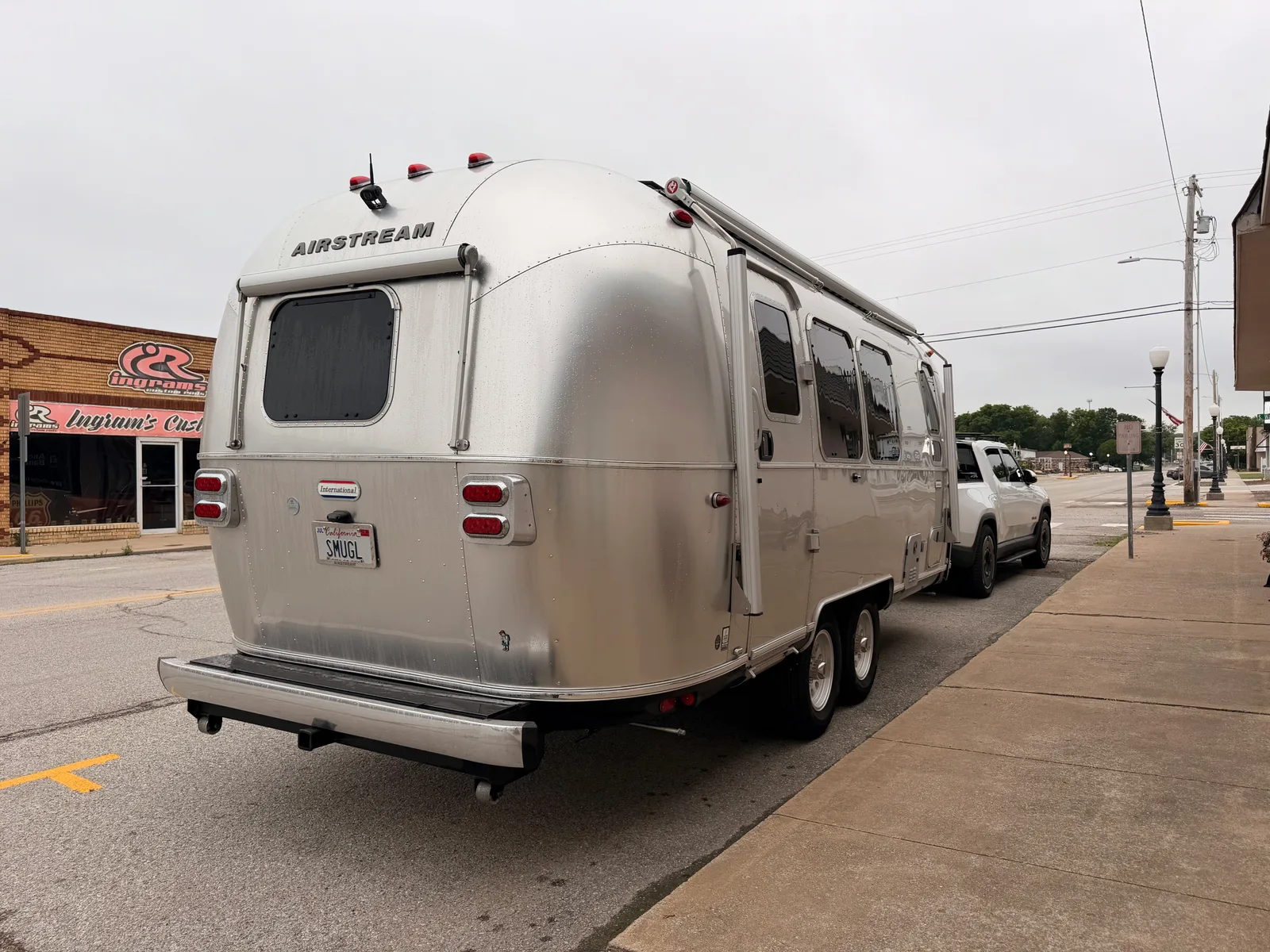 The white Rivian R1T towing the silver Airstream International parked along the brick main street of Locust Grove OK outside Hometown Coffee, low storefronts and a utility pole down the block, overcast morning sky