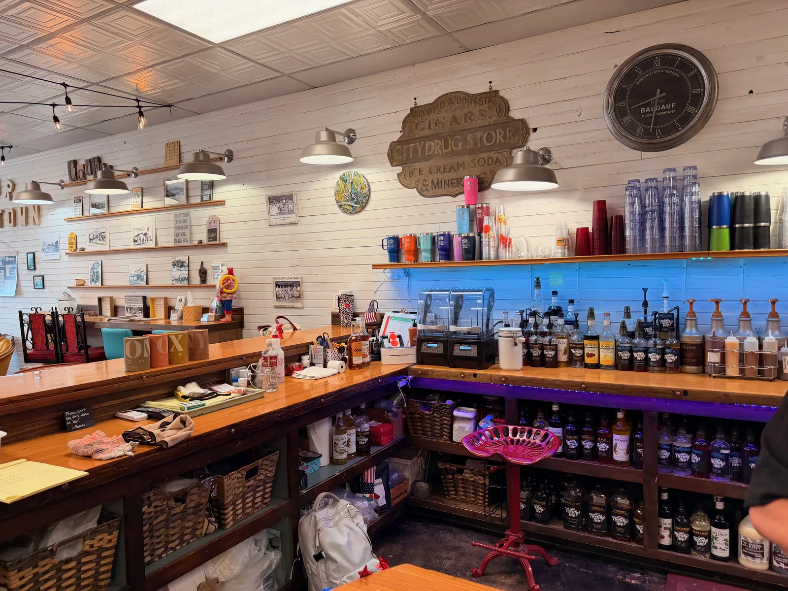 The interior soda-fountain counter at Hometown Coffee in Locust Grove, the original carved "Cigars / City Drug Store / Ice Cream Soda & Mineral Water" sign mounted on the white shiplap wall above a long wooden counter lined with flavored-syrup bottles, a red cast-iron tractor-seat stool, tumblers and tin ceiling above