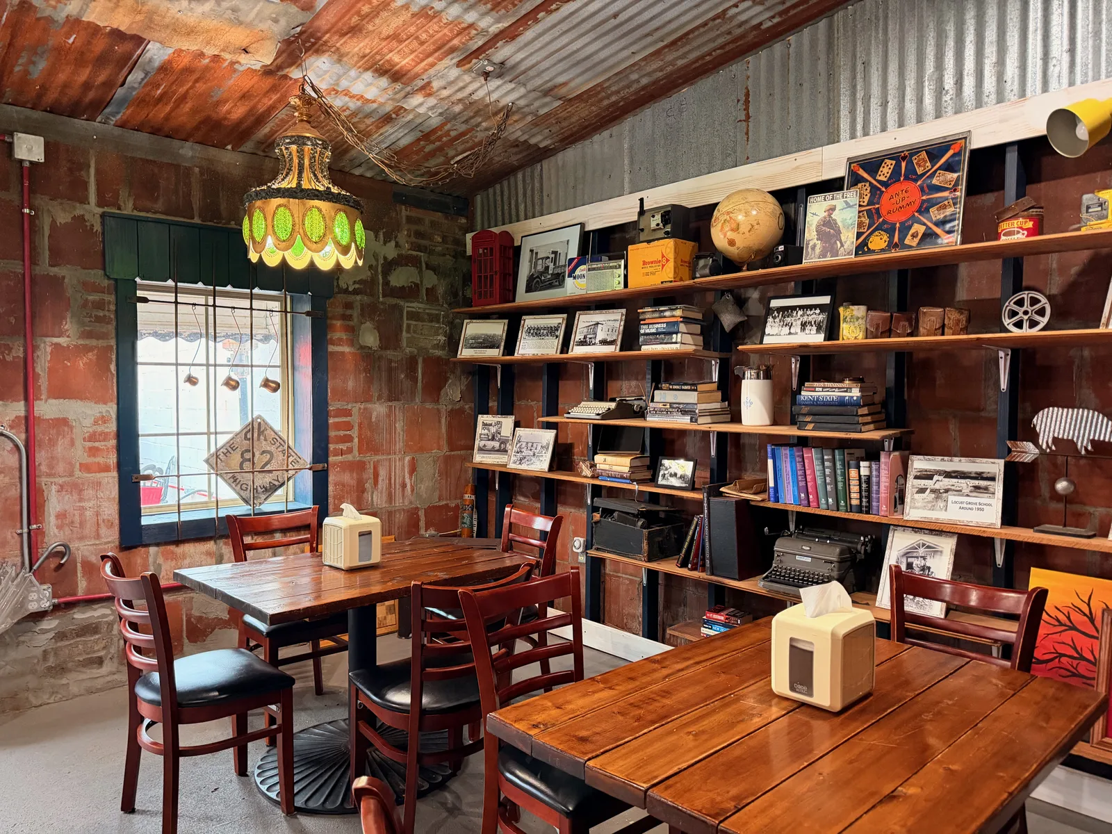 A reading nook inside Hometown Coffee, a tall ladder bookshelf packed with paperbacks, a woven hanging pendant lamp, a wooden table and chairs against an exposed brick wall, a giant red Connect Four set standing beside the shelf