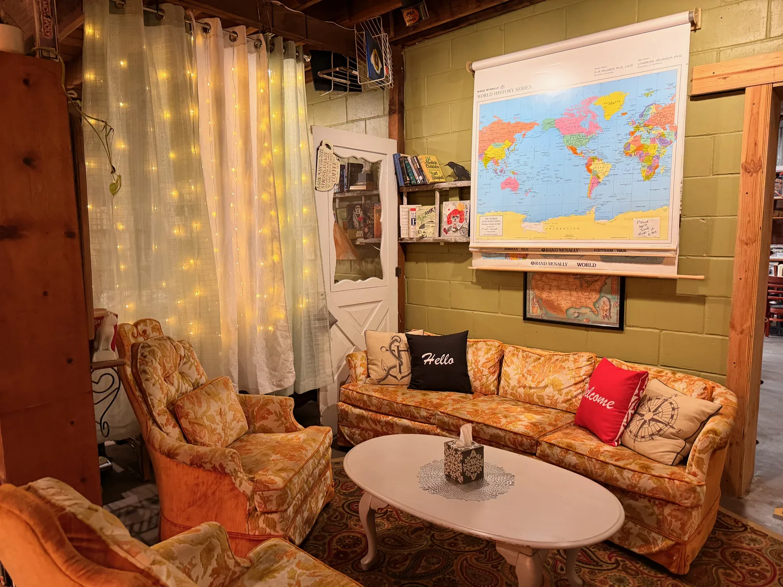 The "Welcome to Our Porch" back lounge at Hometown Coffee, a vintage gold-floral couch with black "Hello" and red "Welcome" throw pillows, a roll-down world map on the green cinder-block wall, string lights overhead, an oval white coffee table on a paisley rug