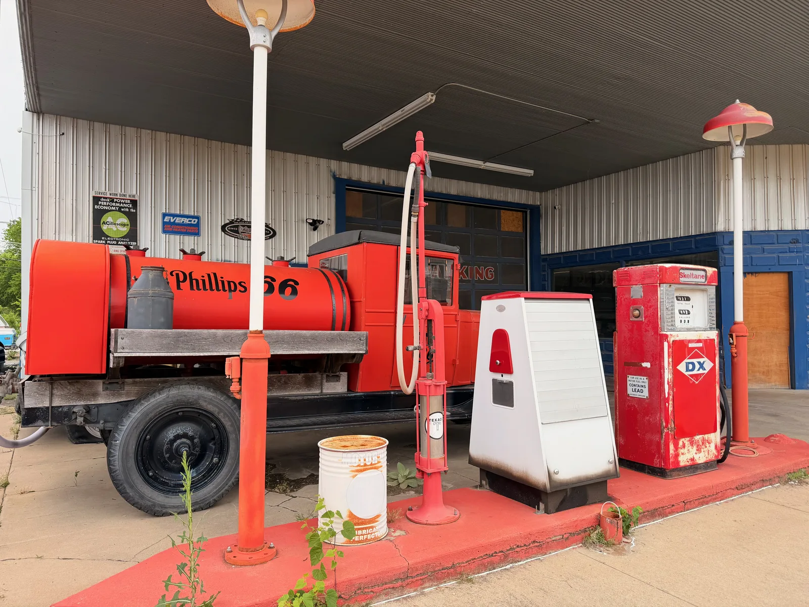A vintage red Phillips 66 tanker truck on display under a metal canopy outside the Locust Grove space, parked beside a tall red visible-register gas pump and a red DX-brand pump, an old oil barrel on the red-painted curb