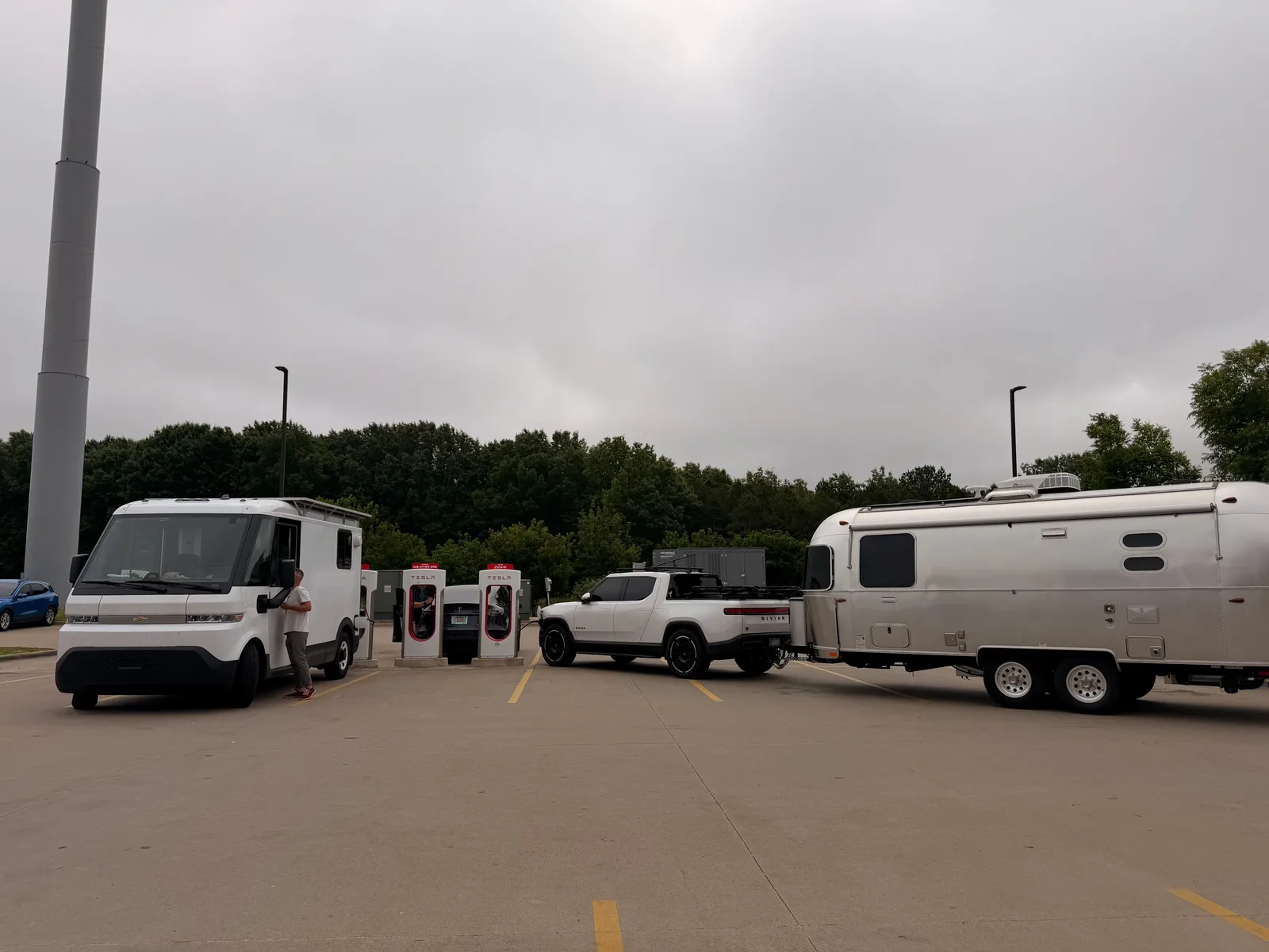 A white GM BrightDrop electric delivery van charging at the Lowell AR Tesla Supercharger right beside the white Rivian R1T and silver Airstream International, red-and-white Supercharger stalls under an overcast NW Arkansas sky