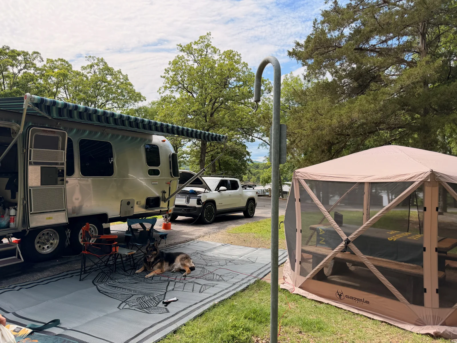 The Airstream, the Rivian R1T, and a screened gazebo arranged on the Indian Point campsite among the trees, camp chairs and gear out, late-afternoon Ozark light