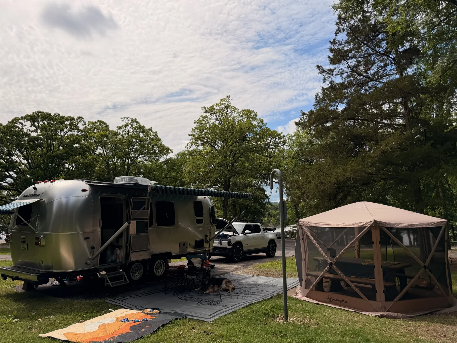 The silver Airstream International set up beside a screened gazebo tent on the wooded lakeside campsite at Indian Point on Table Rock Lake, gravel pad and tall trees, the white Rivian R1T alongside