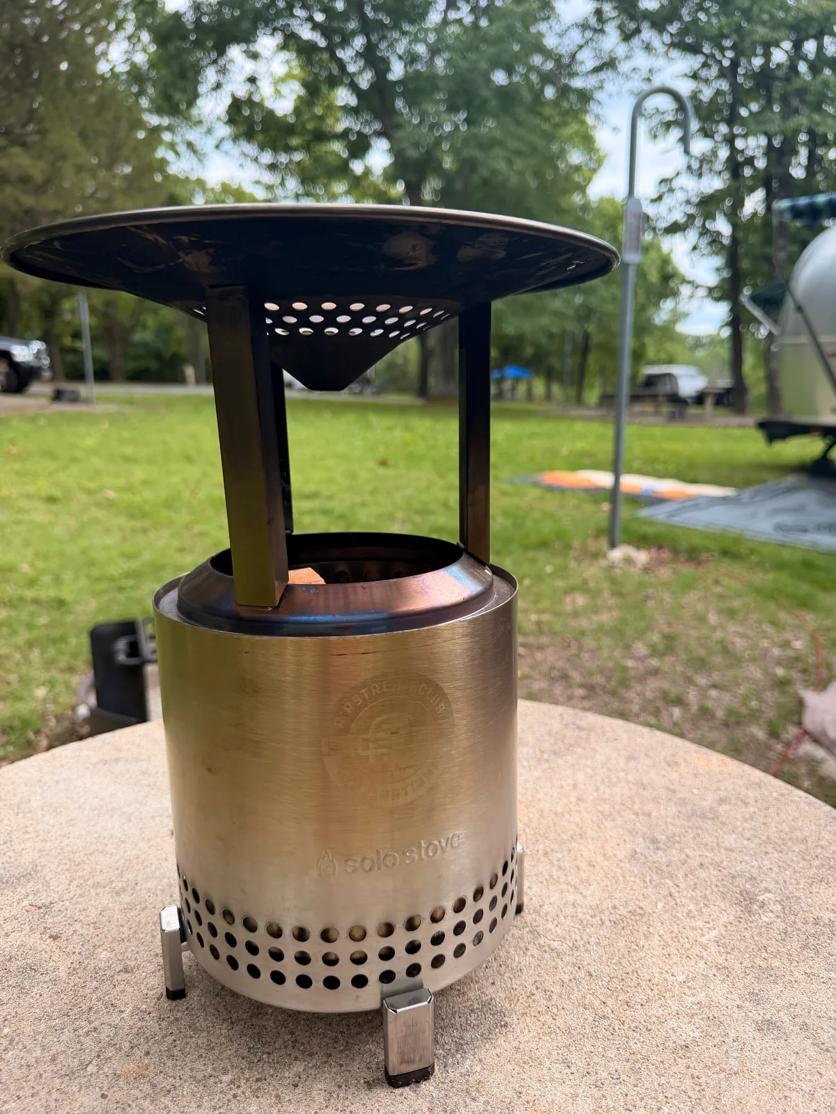 A stainless-steel Solo Stove fire pit standing on the gravel at the Indian Point campsite, trees and a neighboring site behind under evening light