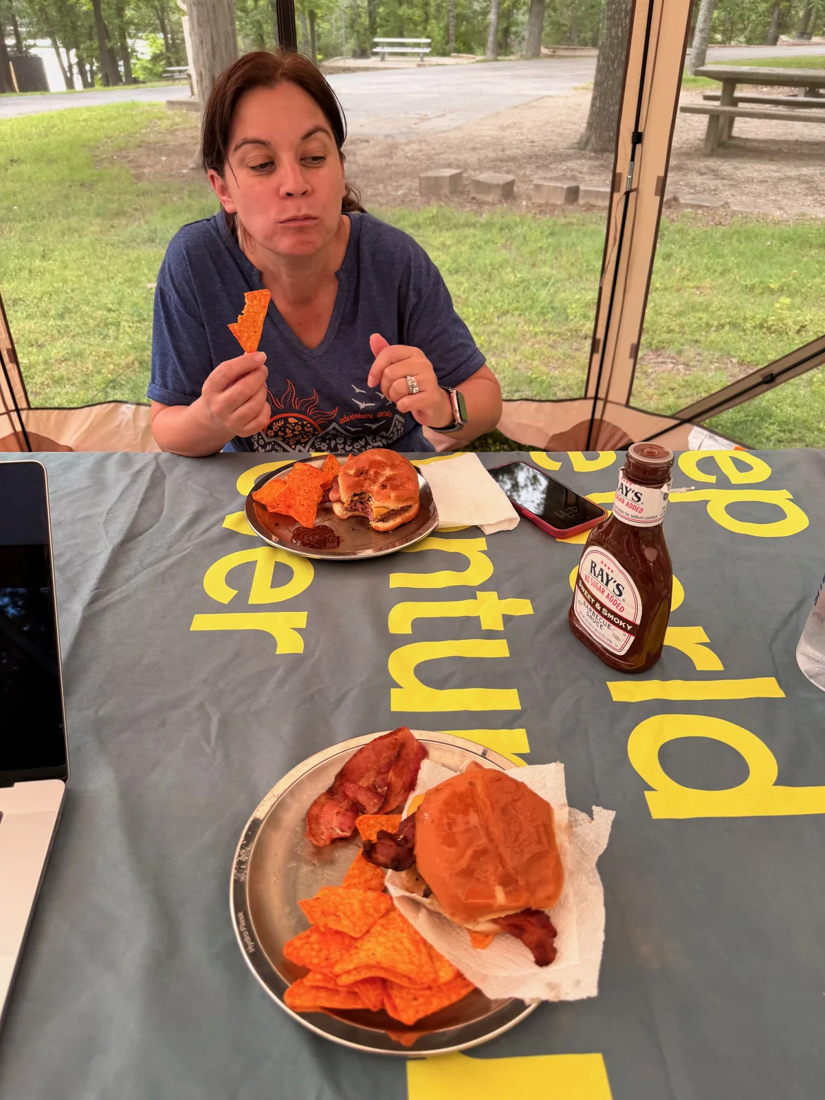 Diana smiling at the camp table under the screened gazebo at Indian Point, a plate of food in front of her on an adventure-print tablecloth