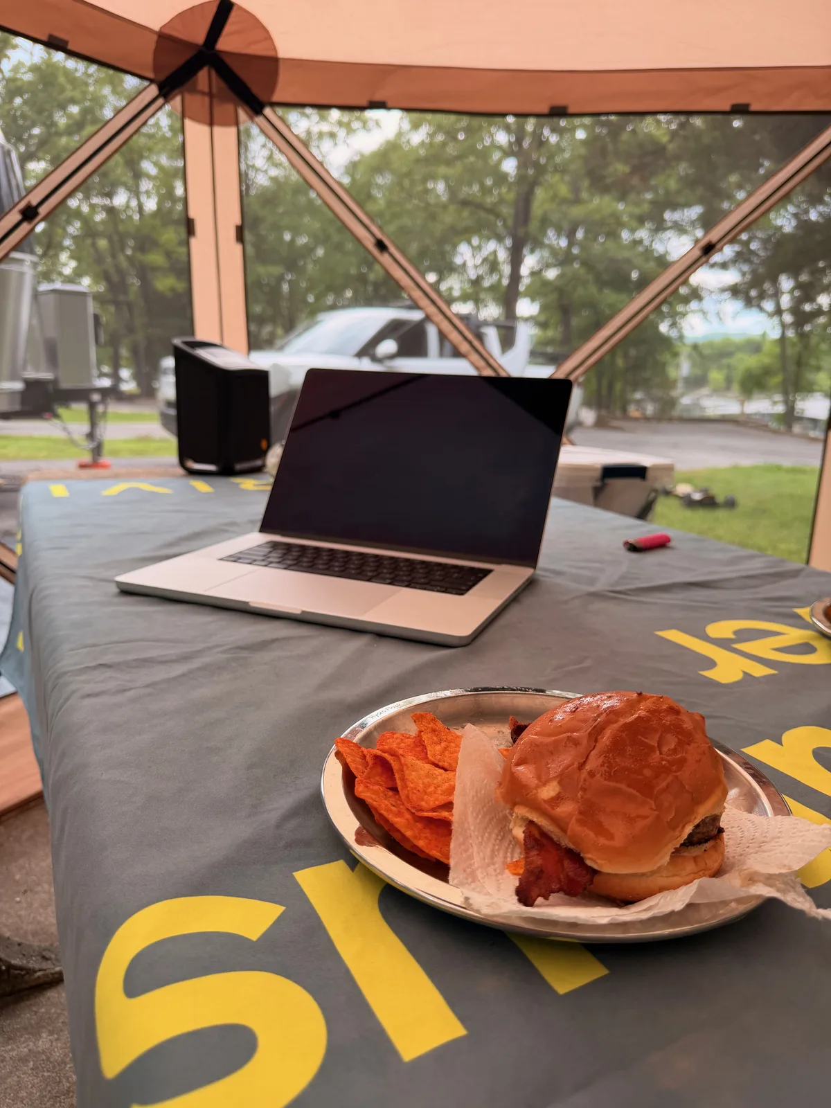 A laptop open beside a plate of barbecue on a camp table under the screened gazebo at Indian Point, the lake and trees beyond the screen