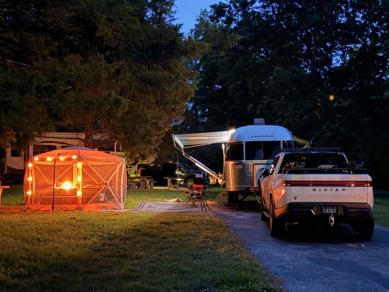 The Indian Point campsite after dark, warm lights glowing on the rig and a neighboring camp under the trees, the white Rivian R1T parked alongside the Airstream