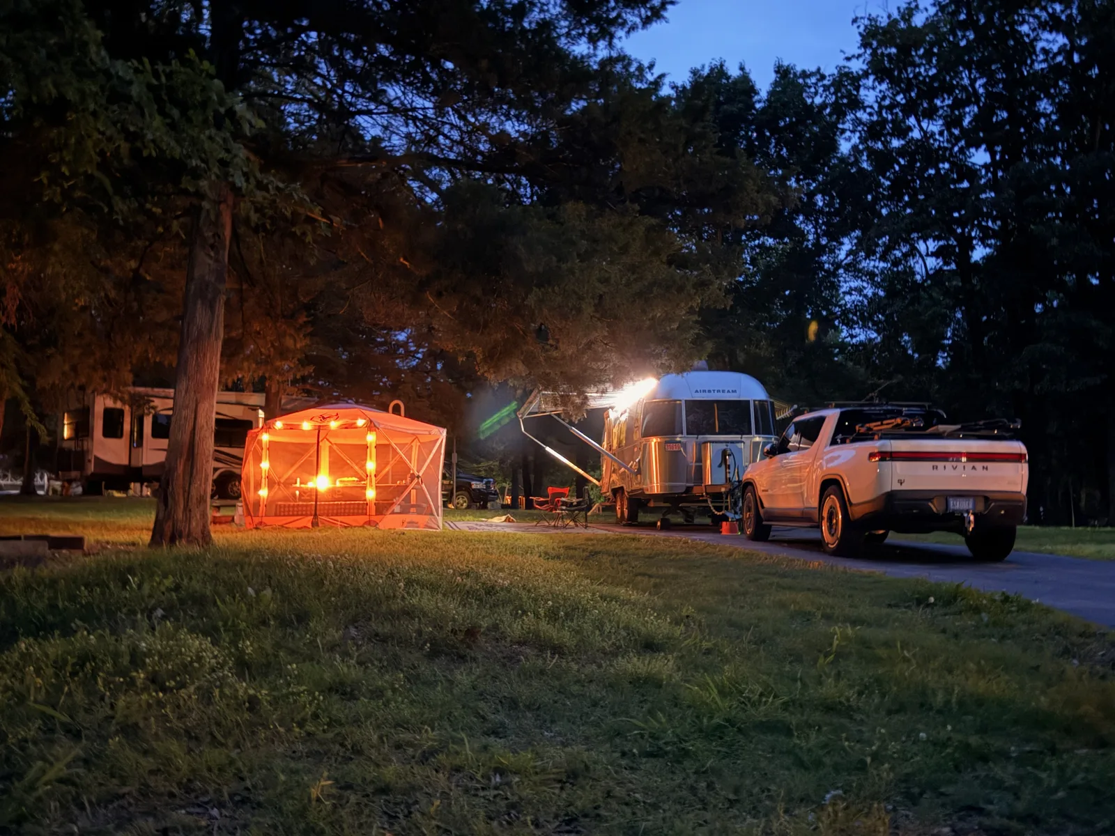 The full Indian Point campsite at blue-hour dusk, the Airstream lit from inside, the Gazelle screen gazebo glowing orange, and the white Rivian R1T parked alongside under the trees