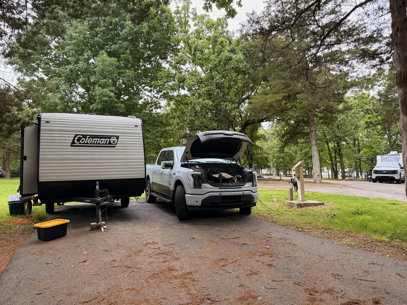 The Indian Point campsite in the morning, a neighboring A-frame pop-up camper and the white Rivian R1T along the wooded campground road, people stirring in the distance